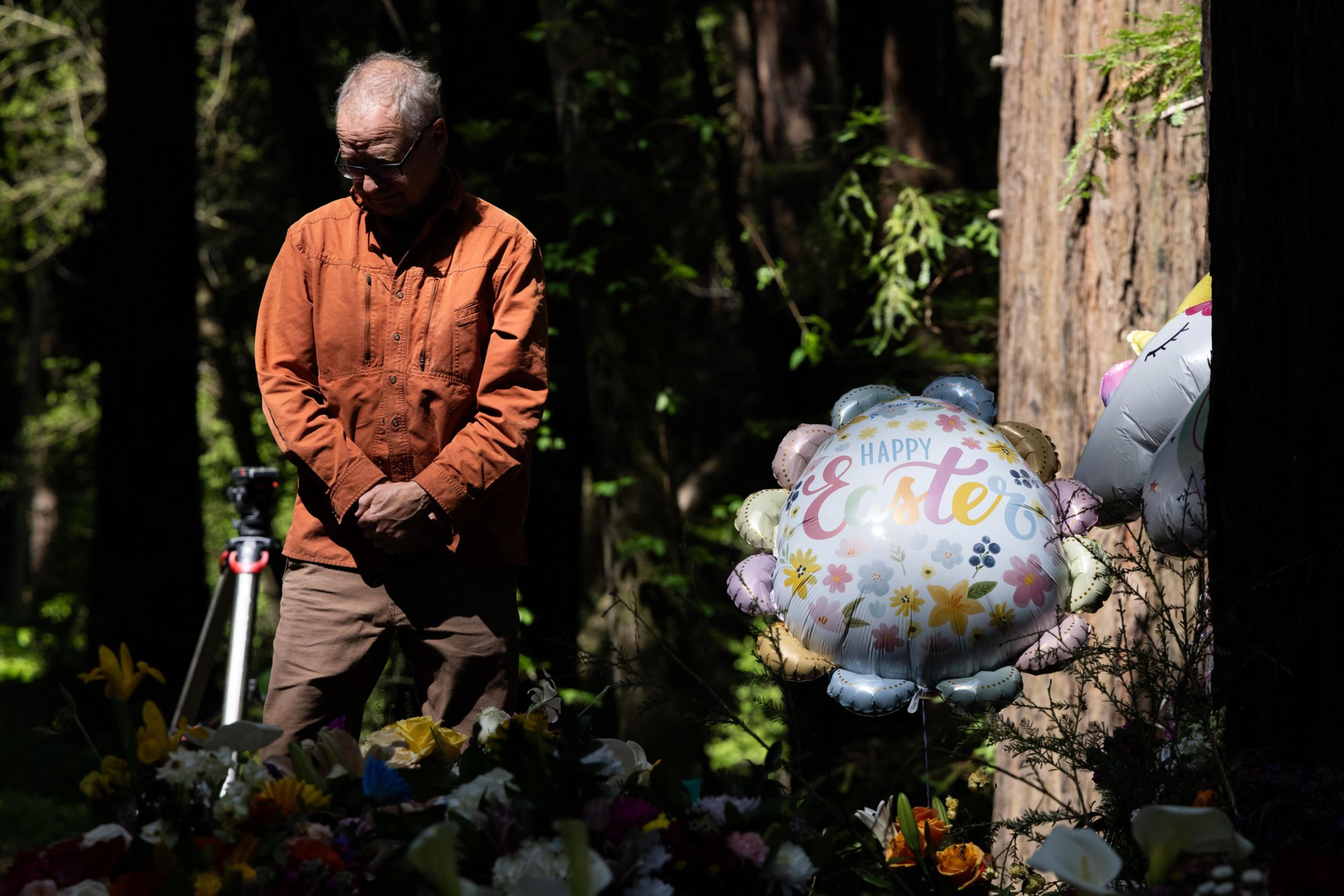 A man in an orange jacket stands solemnly in a wooded area beside an arrangement of flowers and a balloon reading "Happy Easter," with soft sunlight filtering through.