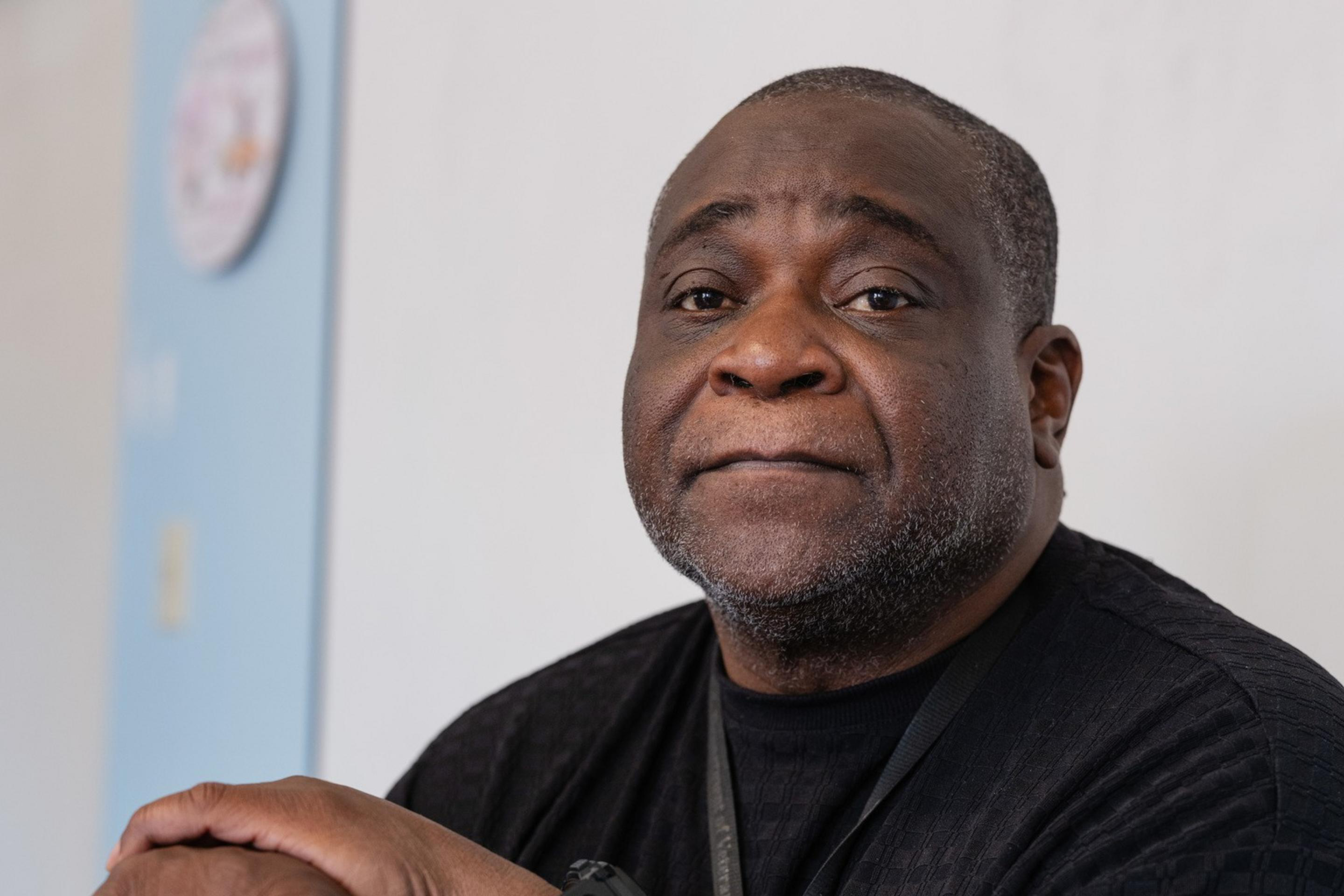 A man sits, resting on a cane, wearing a black shirt and watch. The background shows a light blue and white wall with a round object on it.