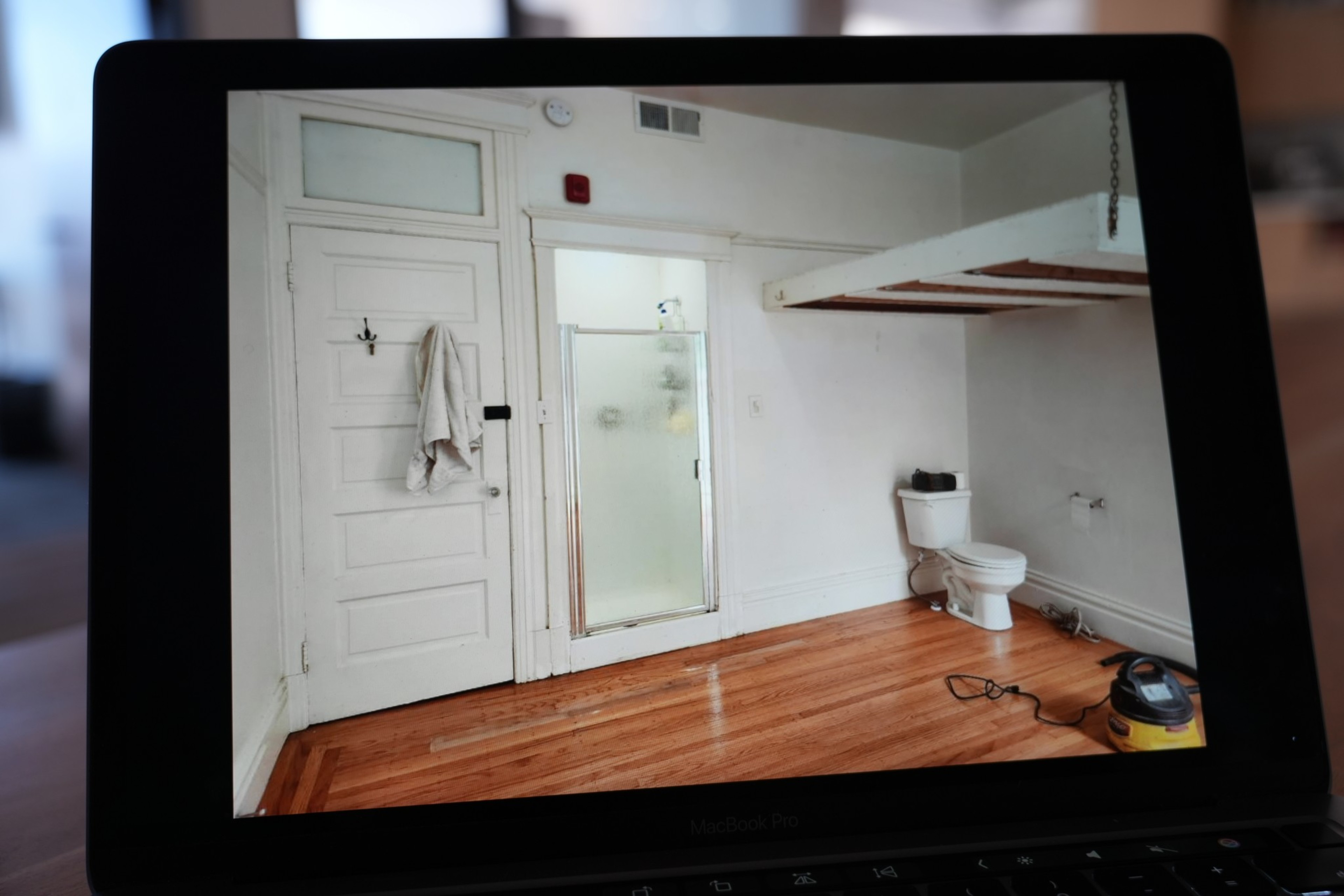 A small room with wooden floors features a white door with a hanging towel, a frosted glass shower, a toilet, and a wet/dry vacuum on the floor.