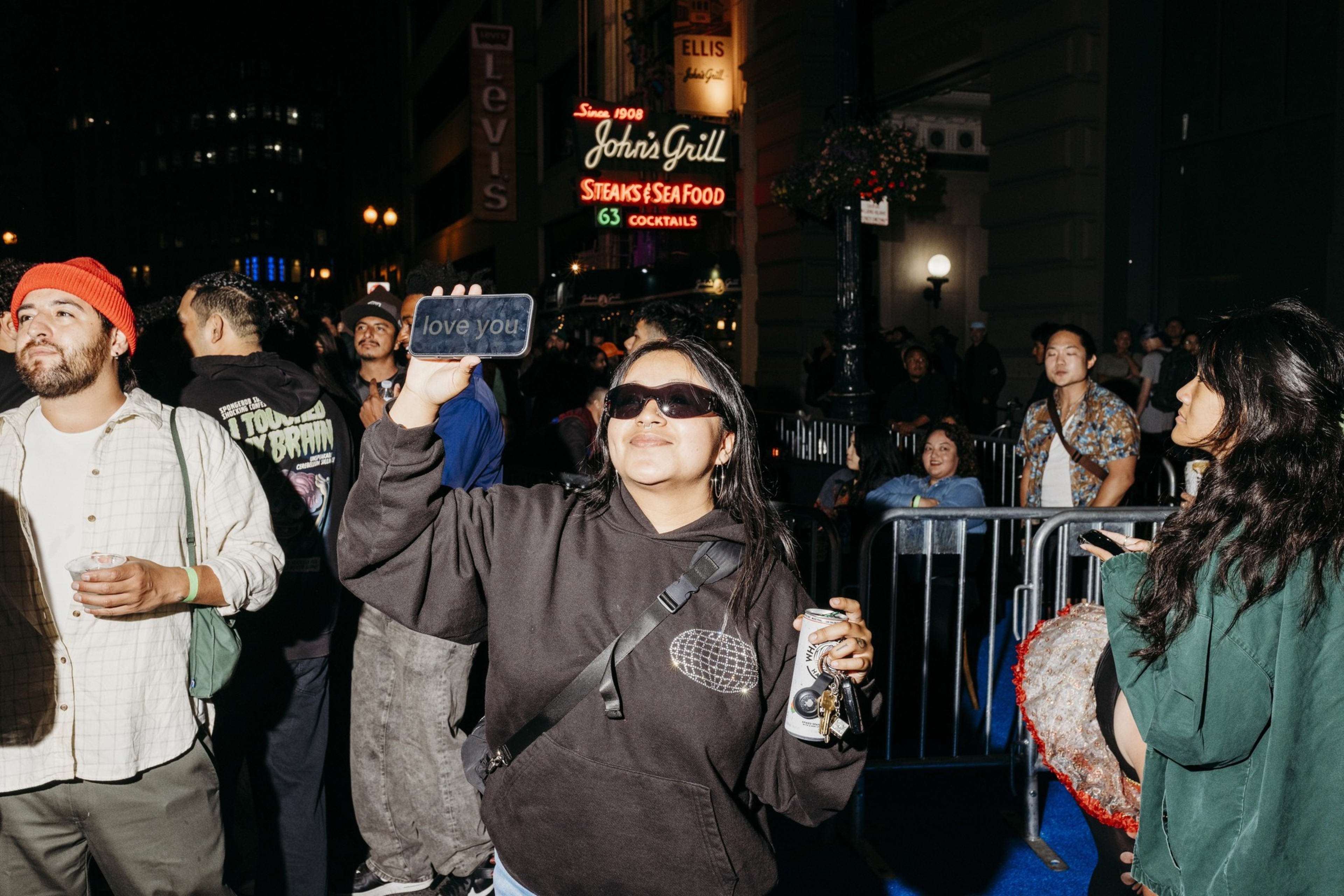 A smiling woman in sunglasses holds a phone displaying “love you” and a can, while people stand around at a nighttime outdoor event near a lit restaurant sign.