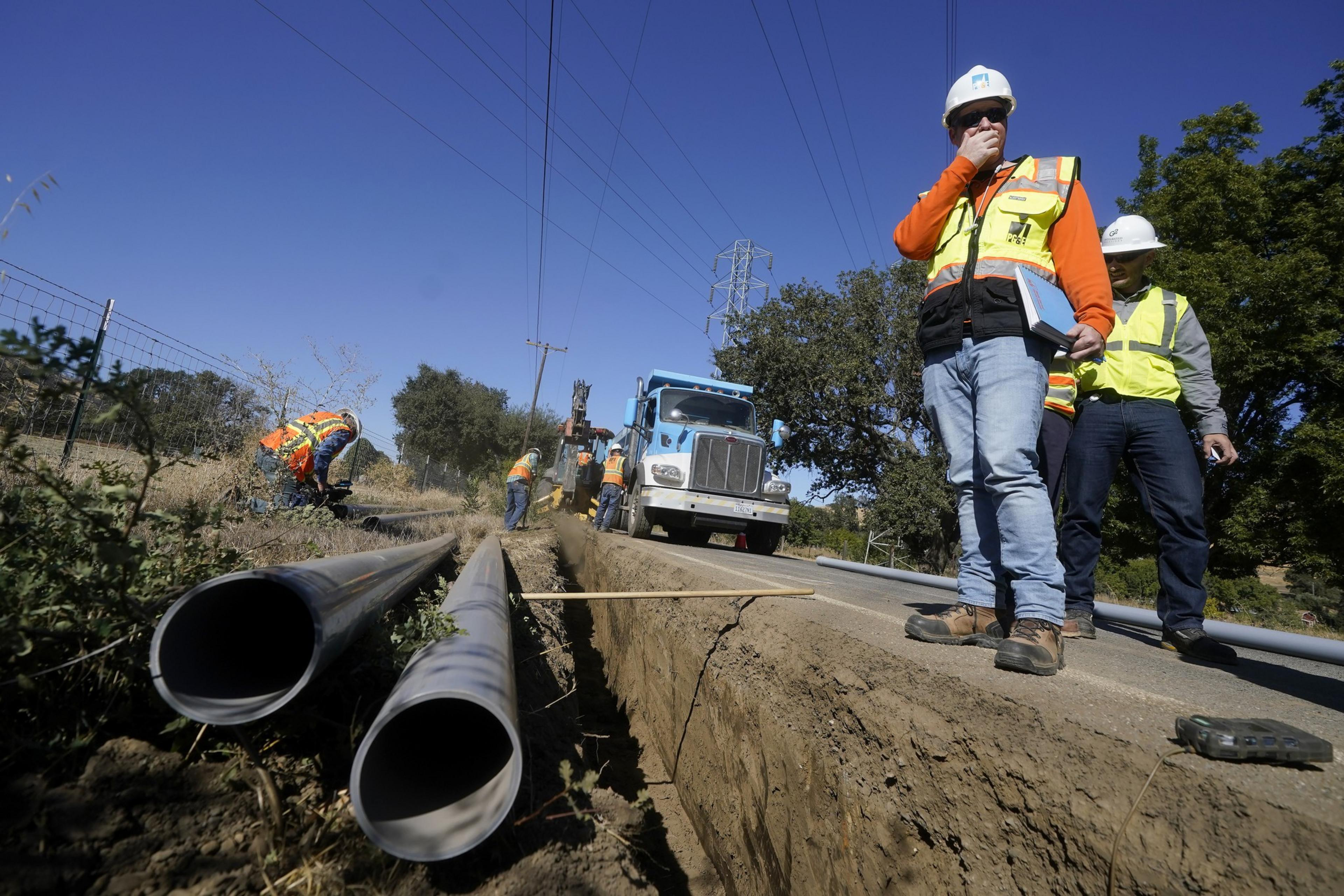 Electric utility workers in orange shirts and neon yellow vests stand beside metal pipes on the ground.