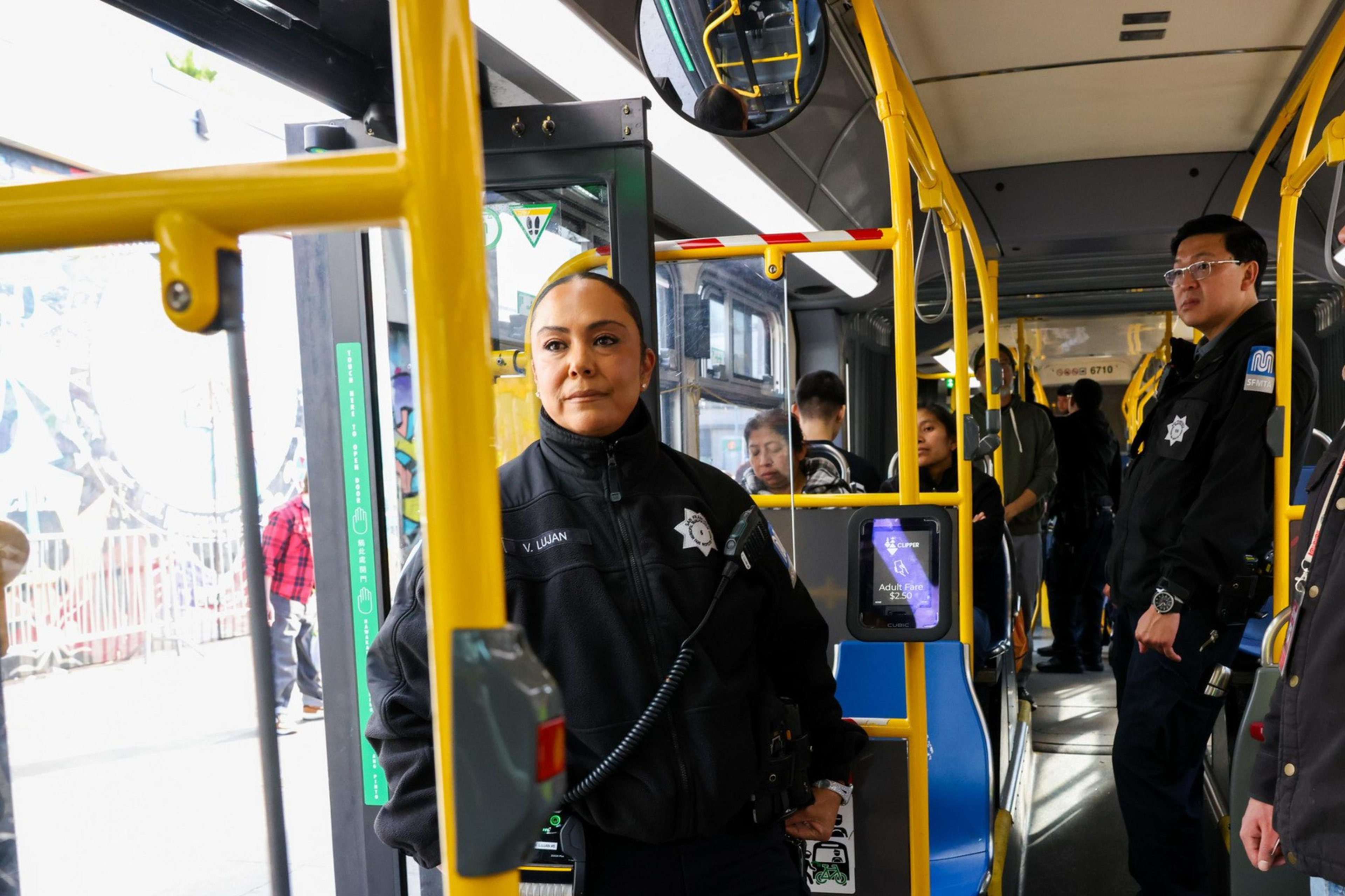 A female officer in uniform stands near a bus door, with another officer and a man in a reflective vest inside. Passengers sit and stand throughout the bus.