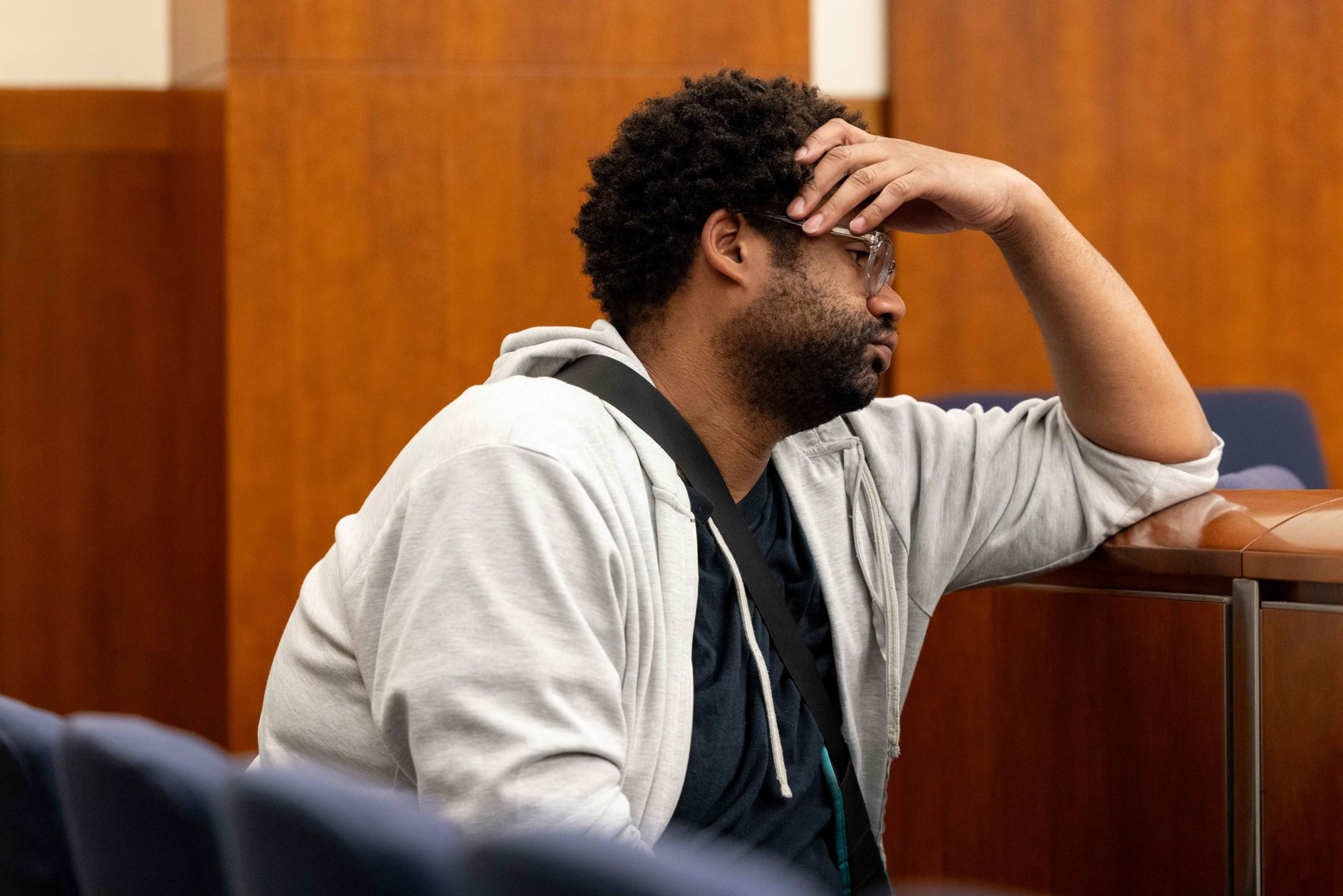 A man with glasses and a beard wearing a gray hoodie leans on a wooden desk with a hand on his forehead, appearing deep in thought or stressed.