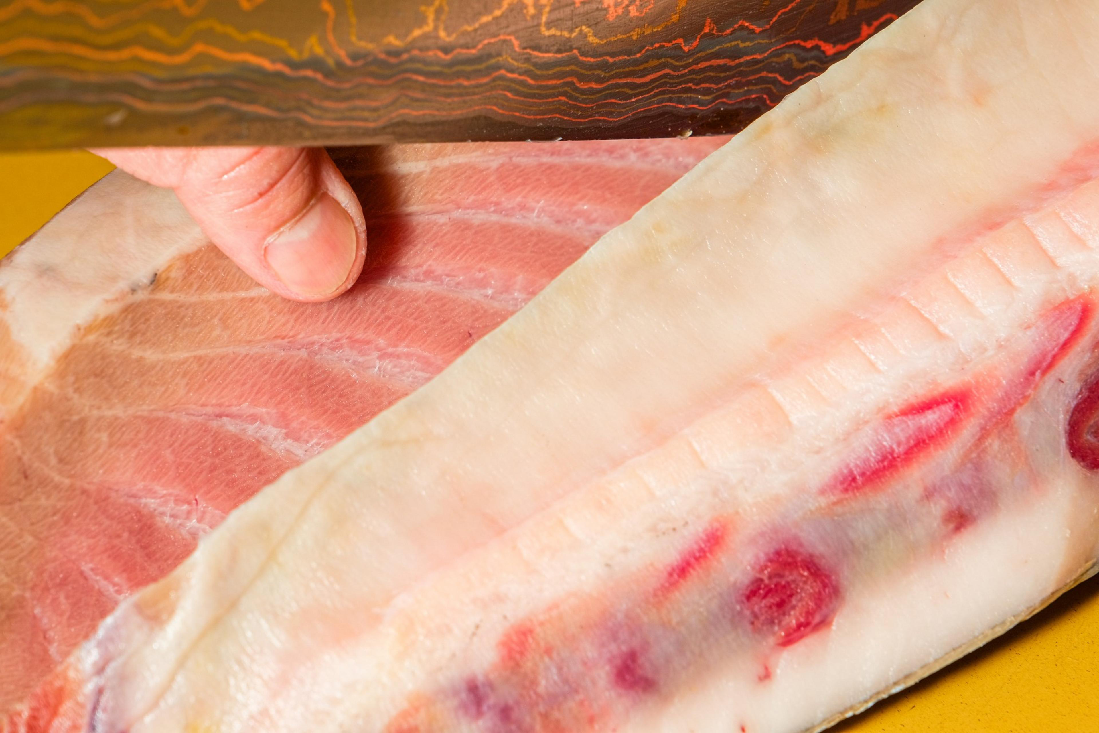 A hand uses a large, decorative knife to cut a thick piece of raw meat with a layer of fat and some red patches, likely prepared for cooking.