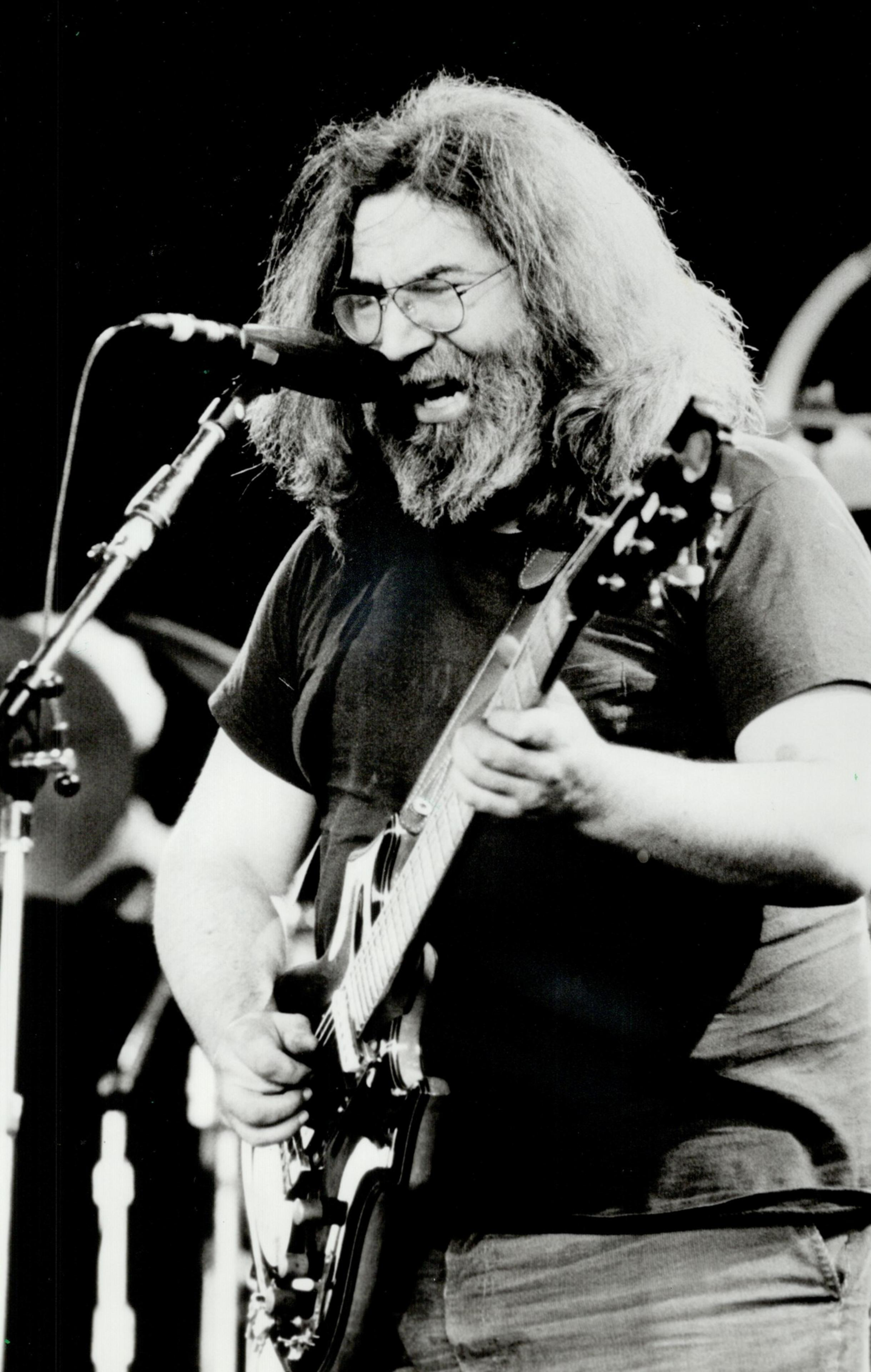 A bearded man with long hair plays an electric guitar passionately, facing a microphone. He's wearing a dark shirt, and the scene is in black and white.