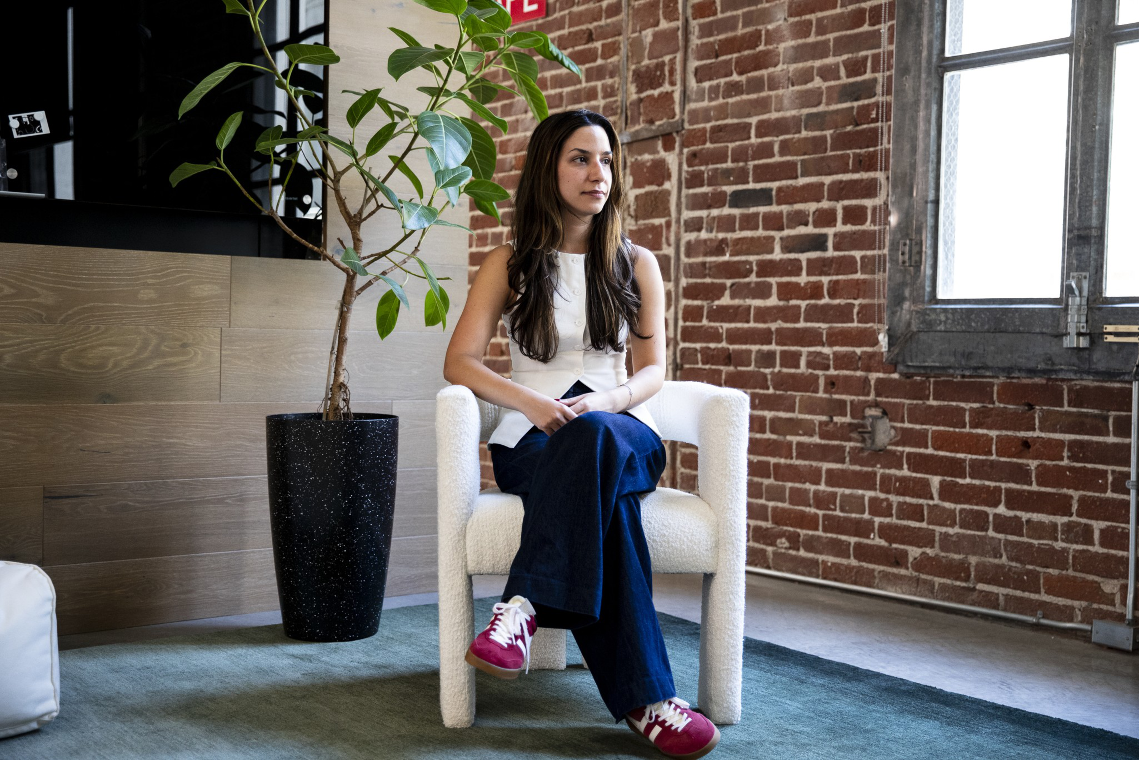 A woman with long brown hair sits cross-legged in a white chair, wearing a white top, dark blue pants, and red sneakers, against a brick wall background.