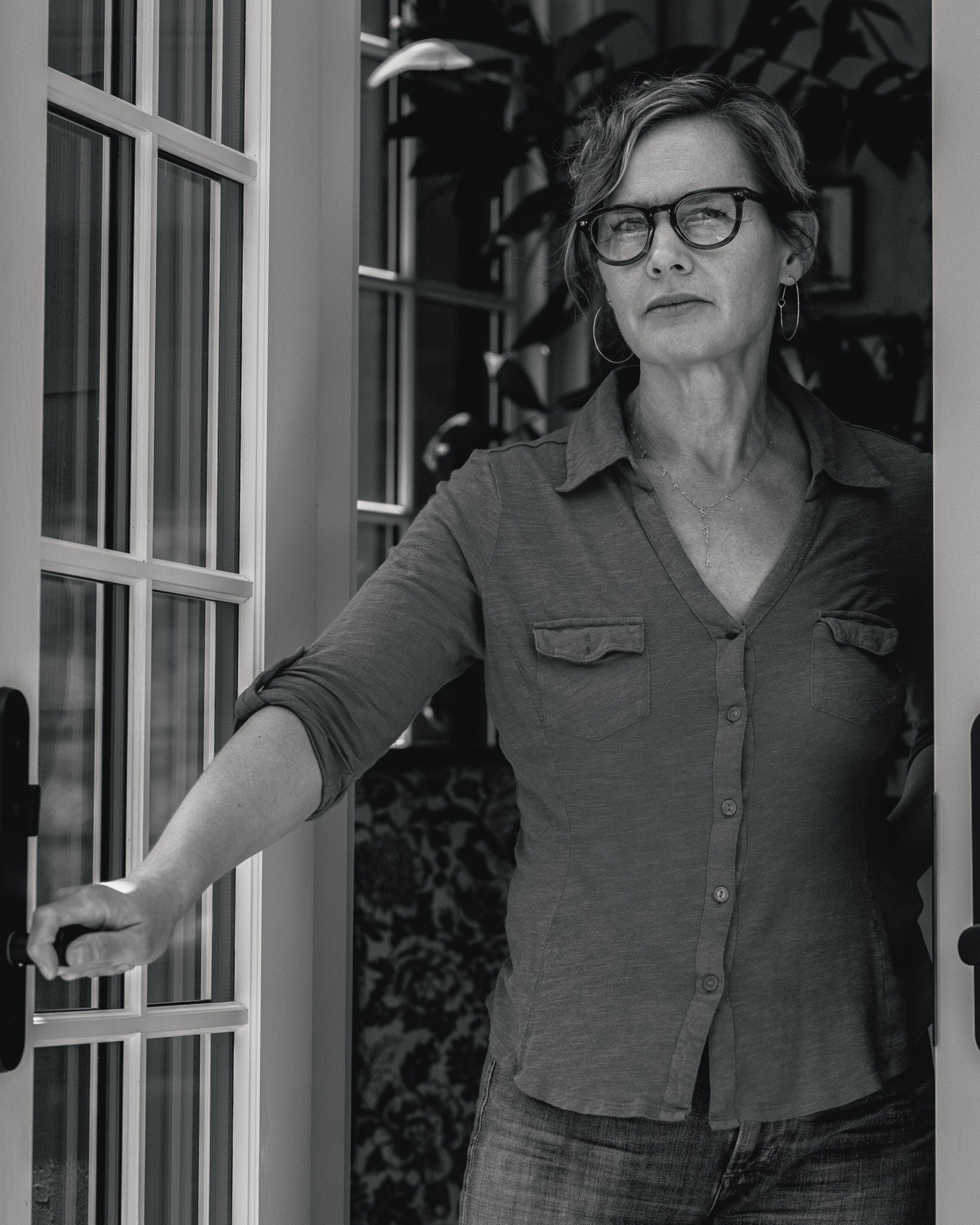 A woman wearing glasses and a casual button-up shirt stands in an open doorway, holding the door handle, with foliage visible in the background.