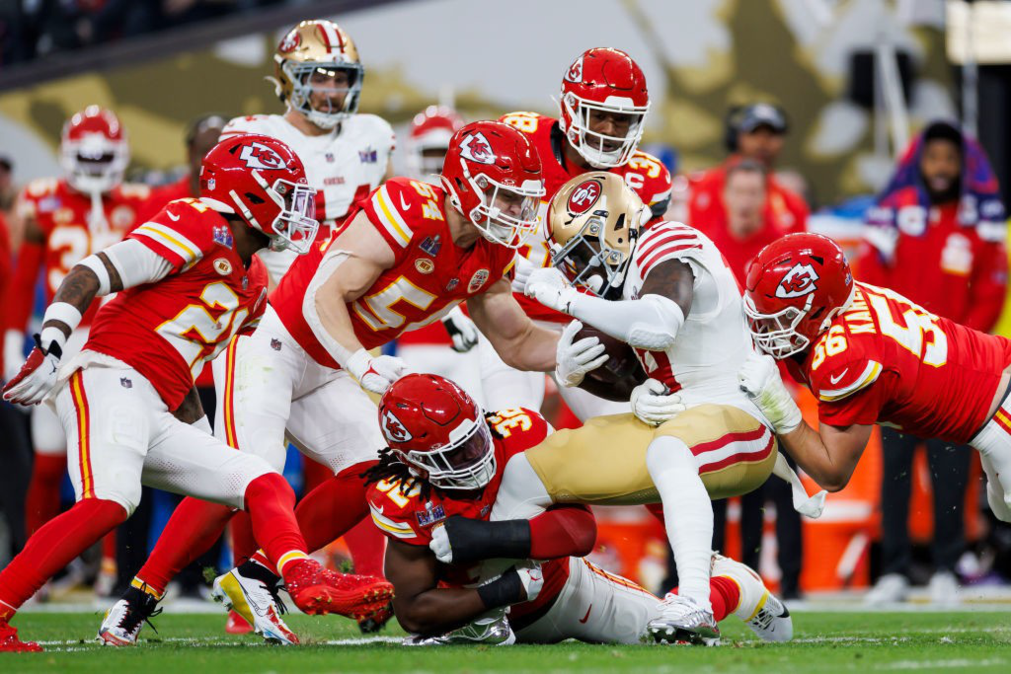 Players in red and white football uniforms engage in a tackle. The player in white is being brought down by multiple players in red jerseys on a grassy field.
