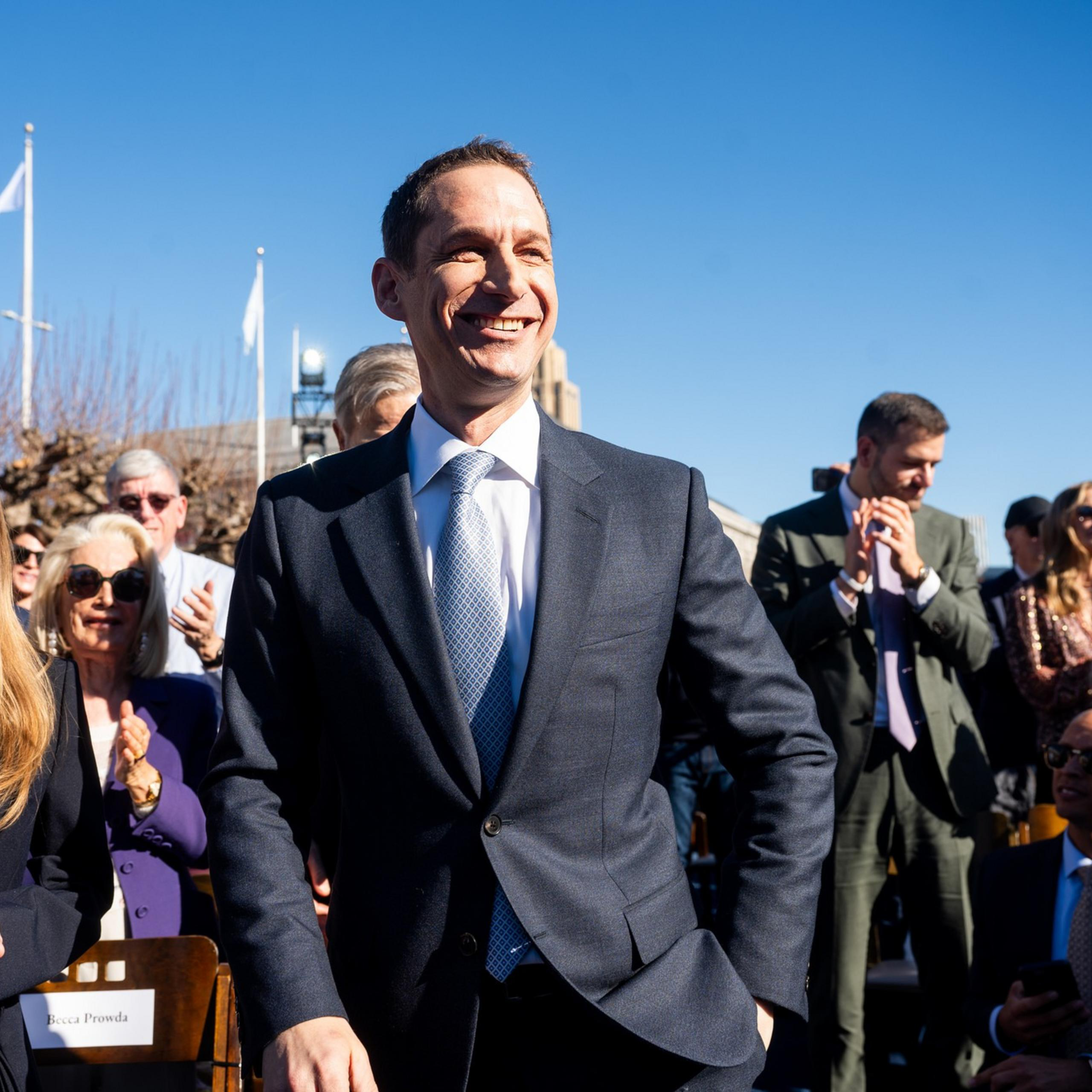A man in a suit smiles broadly among a crowd outdoors. People are clapping, and there's a clear blue sky overhead. A woman with long hair is next to him.