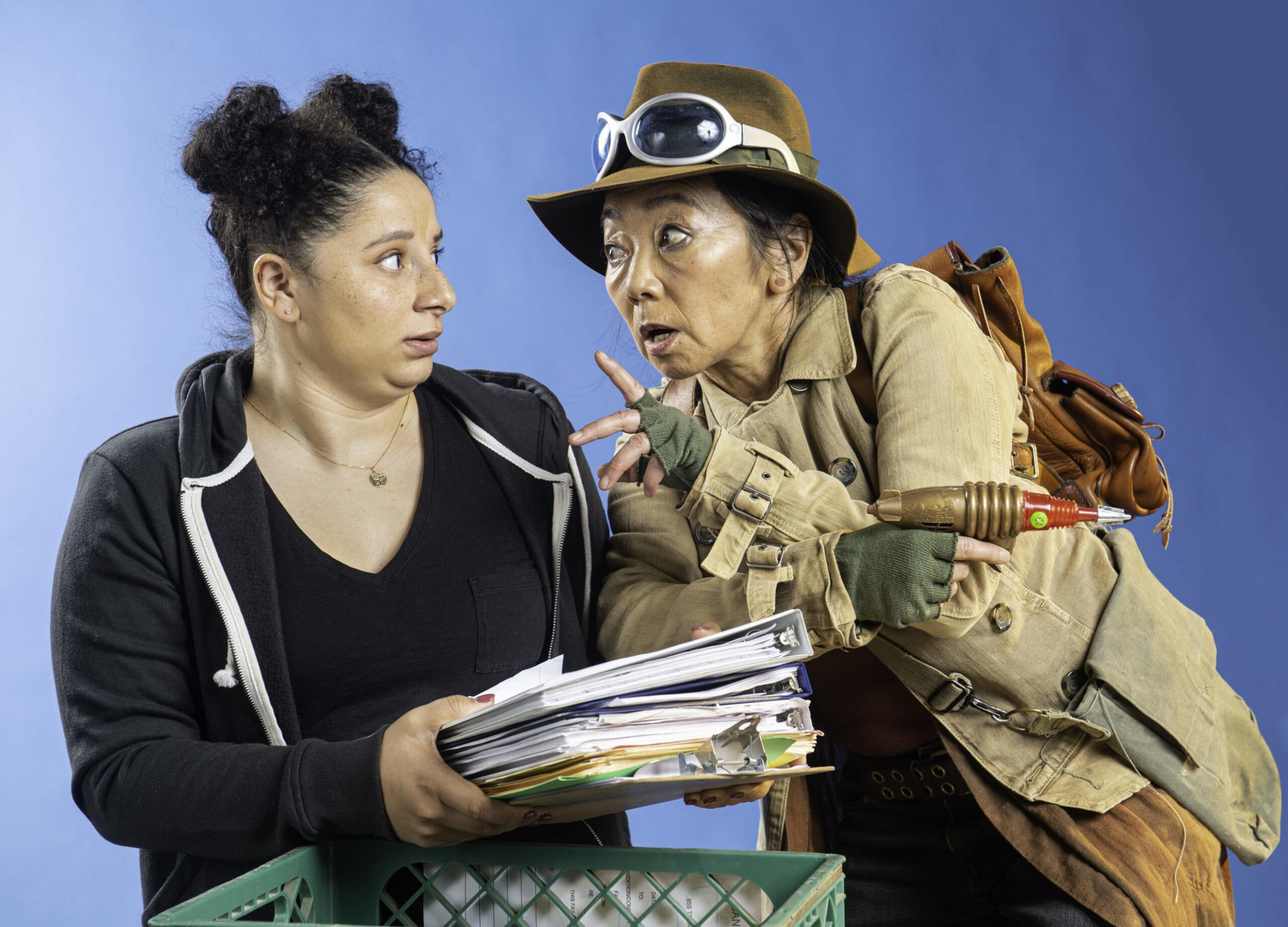 A woman holding a large stack of papers looks surprised as another woman in explorer gear points a toy ray gun at her.