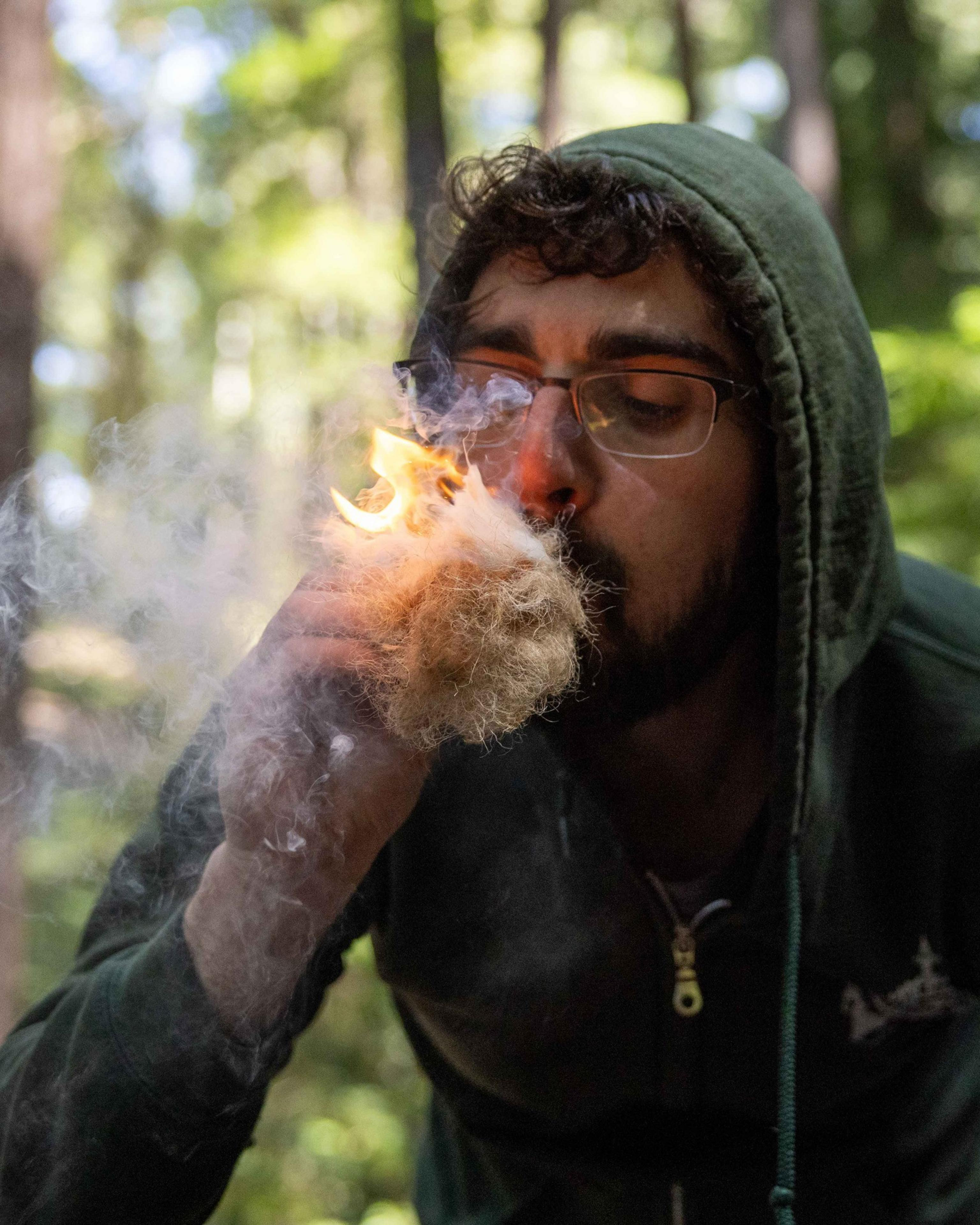 A person in a green hoodie is trying to light dry grass or tinder with smoke and a small flame in a forest setting.