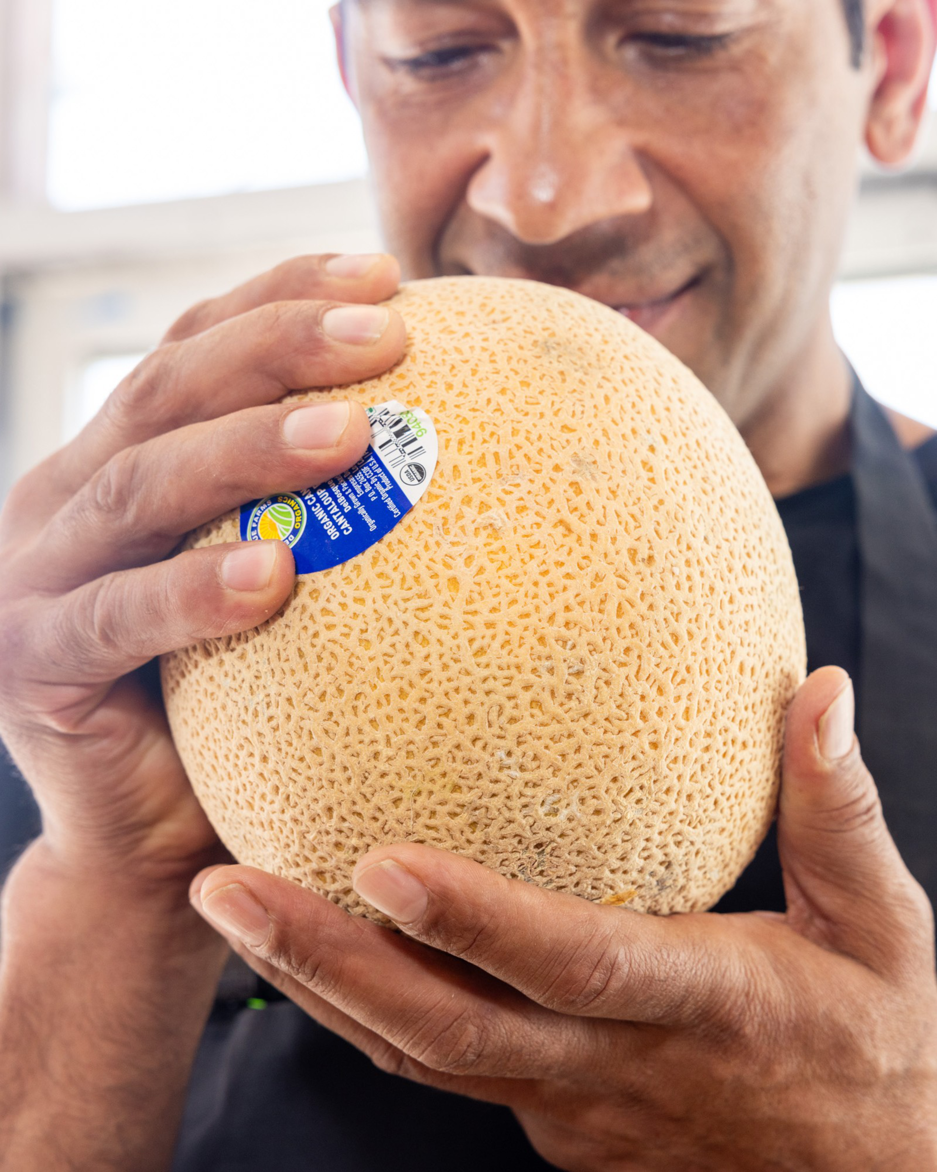 A person is closely inspecting a cantaloupe, holding it gently with both hands. The cantaloupe has a barcode sticker on it, and the person appears focused.