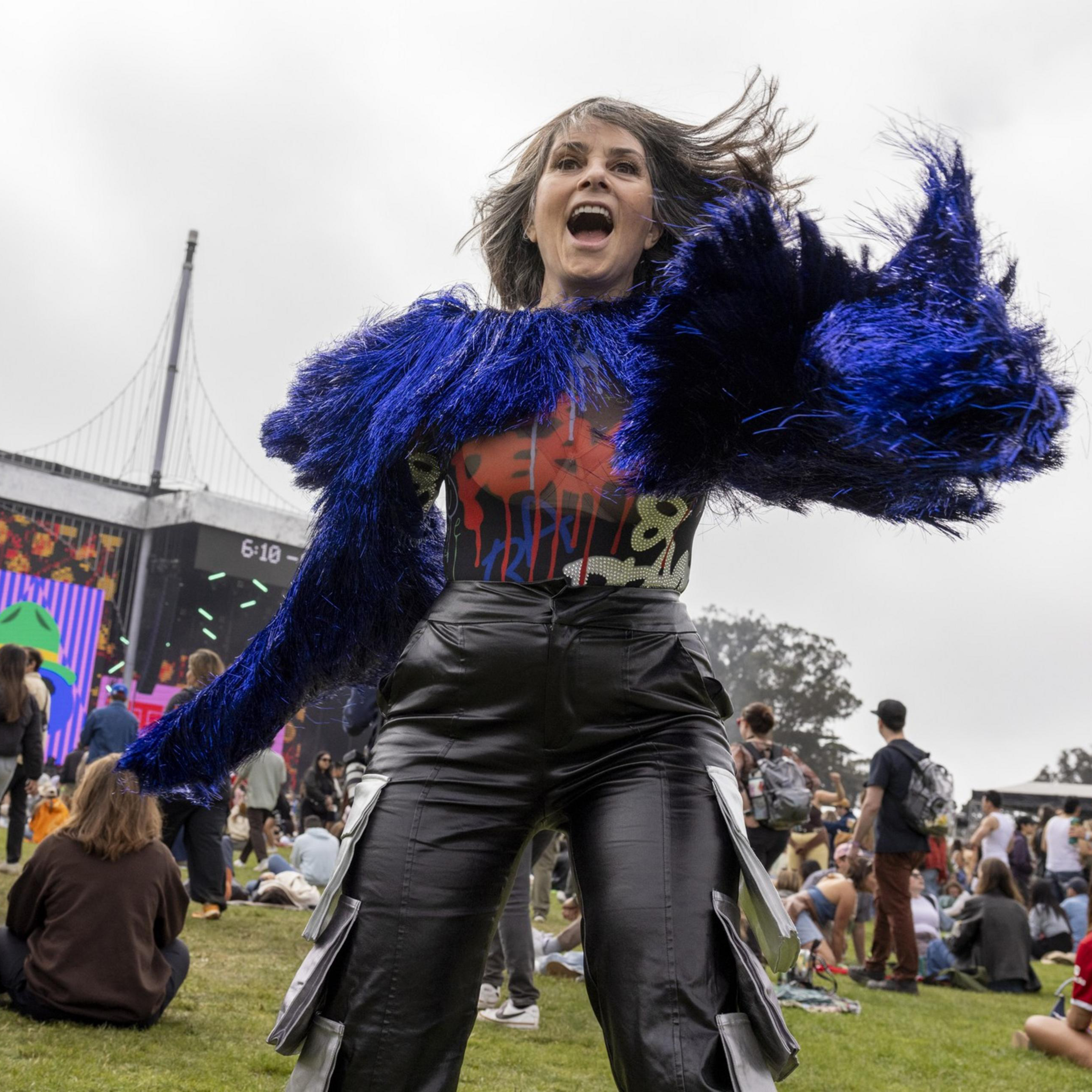 A woman in a vibrant blue, furry jacket dances energetically at an outdoor music festival. The background shows a large crowd and a lit stage with colorful screens.