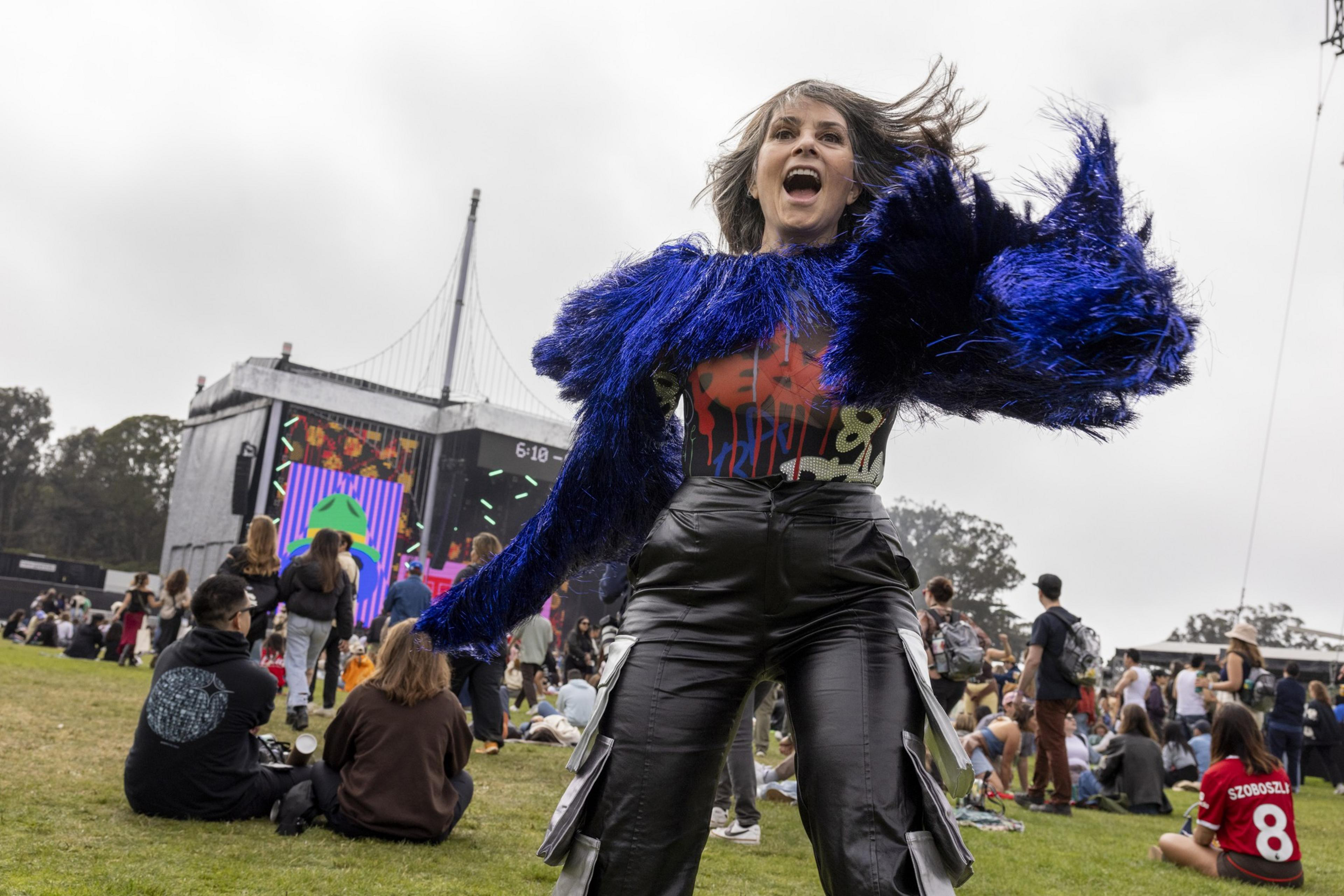 A woman in a vibrant blue, furry jacket dances energetically at an outdoor music festival. The background shows a large crowd and a lit stage with colorful screens.
