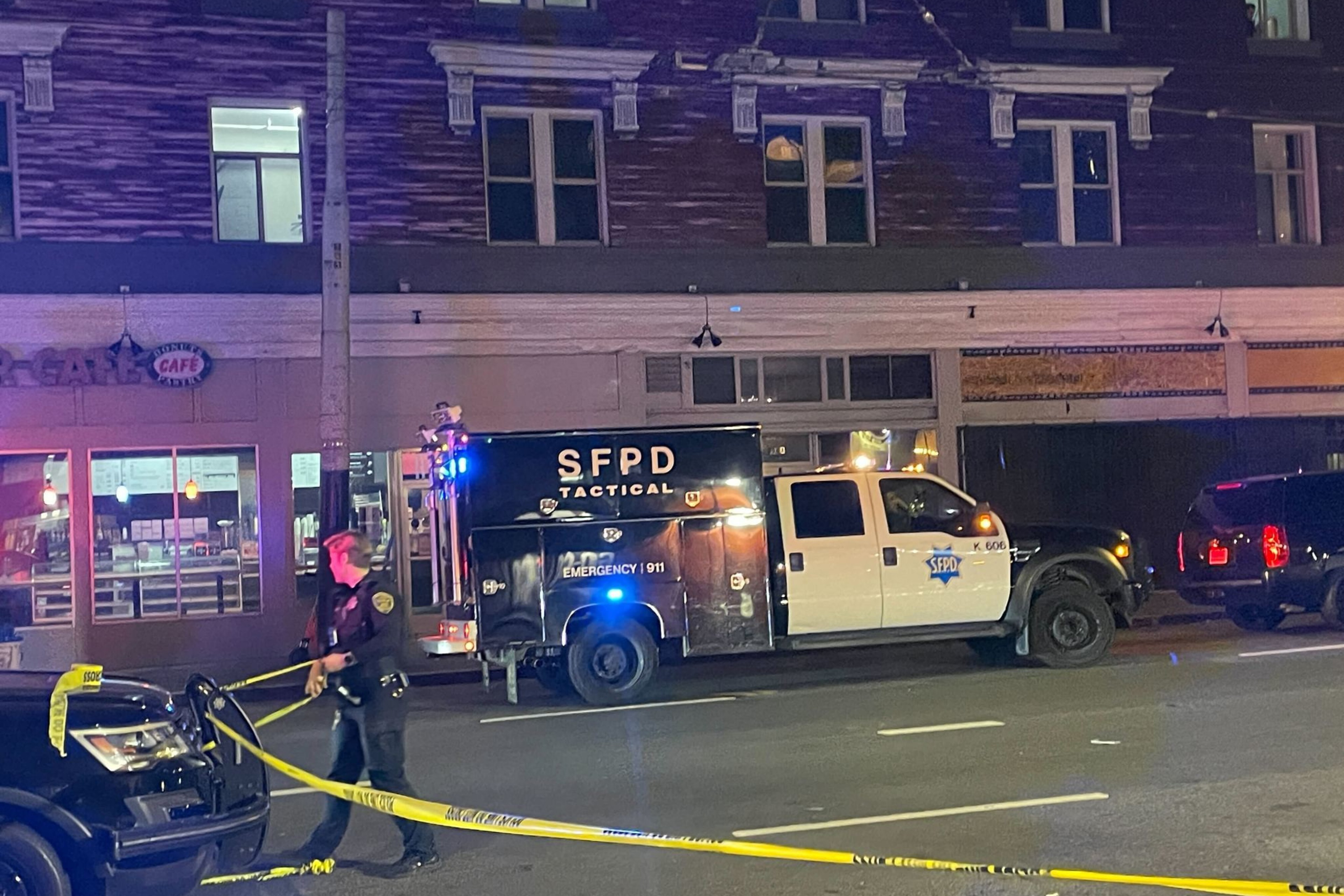 A nighttime scene shows a police tactical vehicle labeled "SFPD" parked on a street. A police officer manages yellow tape, and a building is in the background.