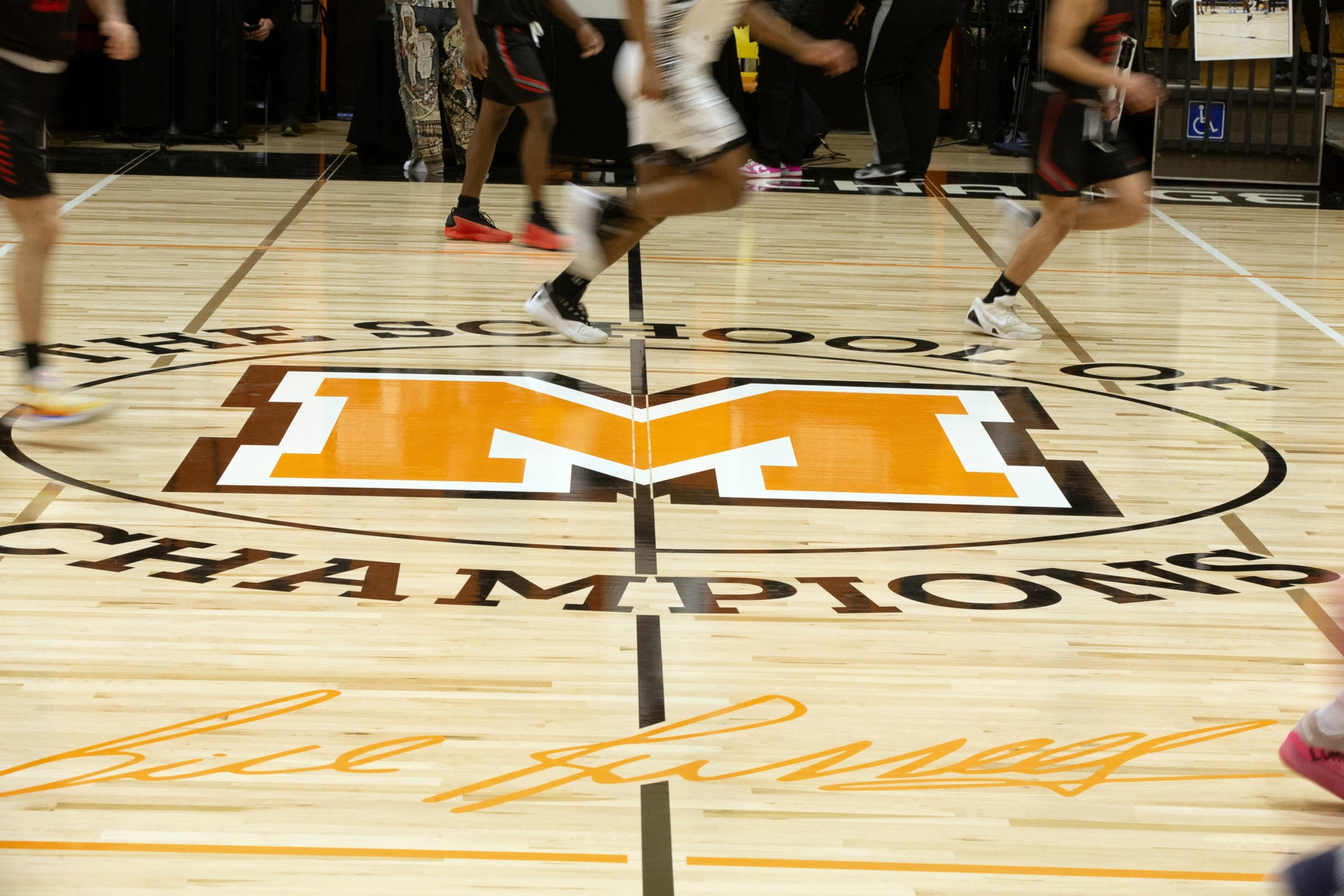 Basketball players run across a court with a large orange &quot;M&quot; and &quot;The School of Champions&quot; written around it, with a signature below.