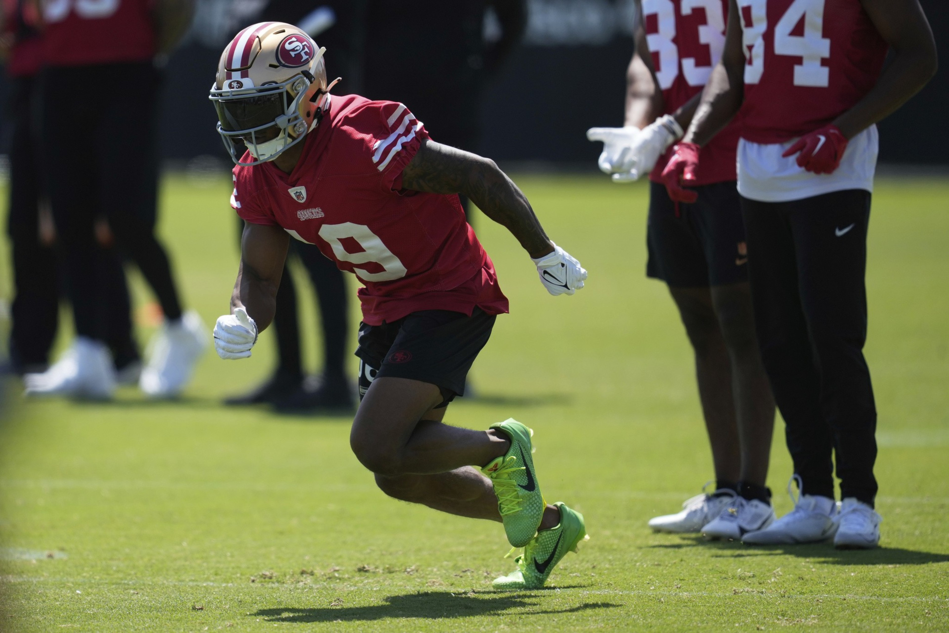 A football player in a red jersey and helmet runs on grass. He wears bright green cleats, and several other players in red jerseys stand nearby.