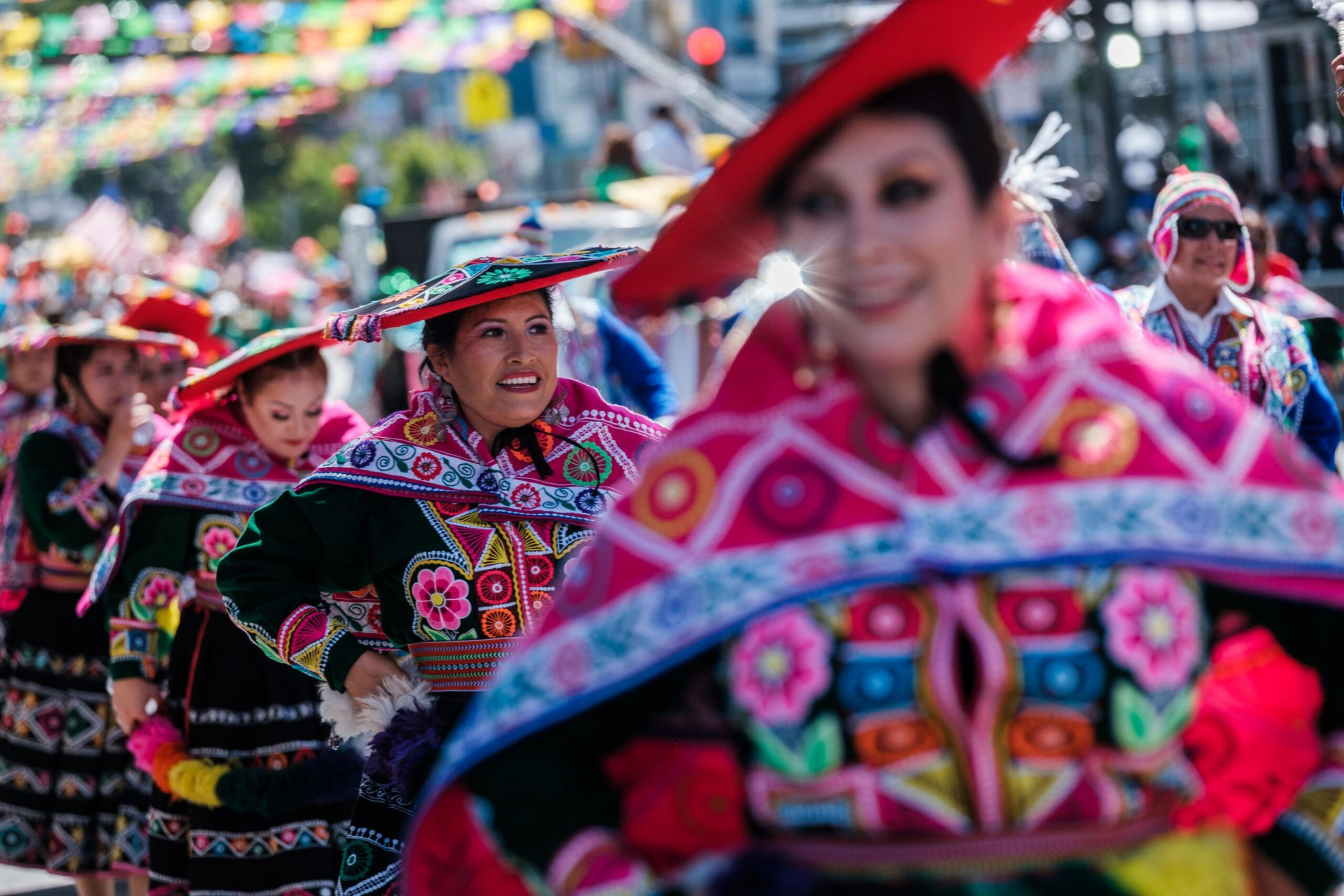 Women wearing brightly colored traditional Andean outfits with elaborate floral embroidery and red hats dance in a festive street parade.