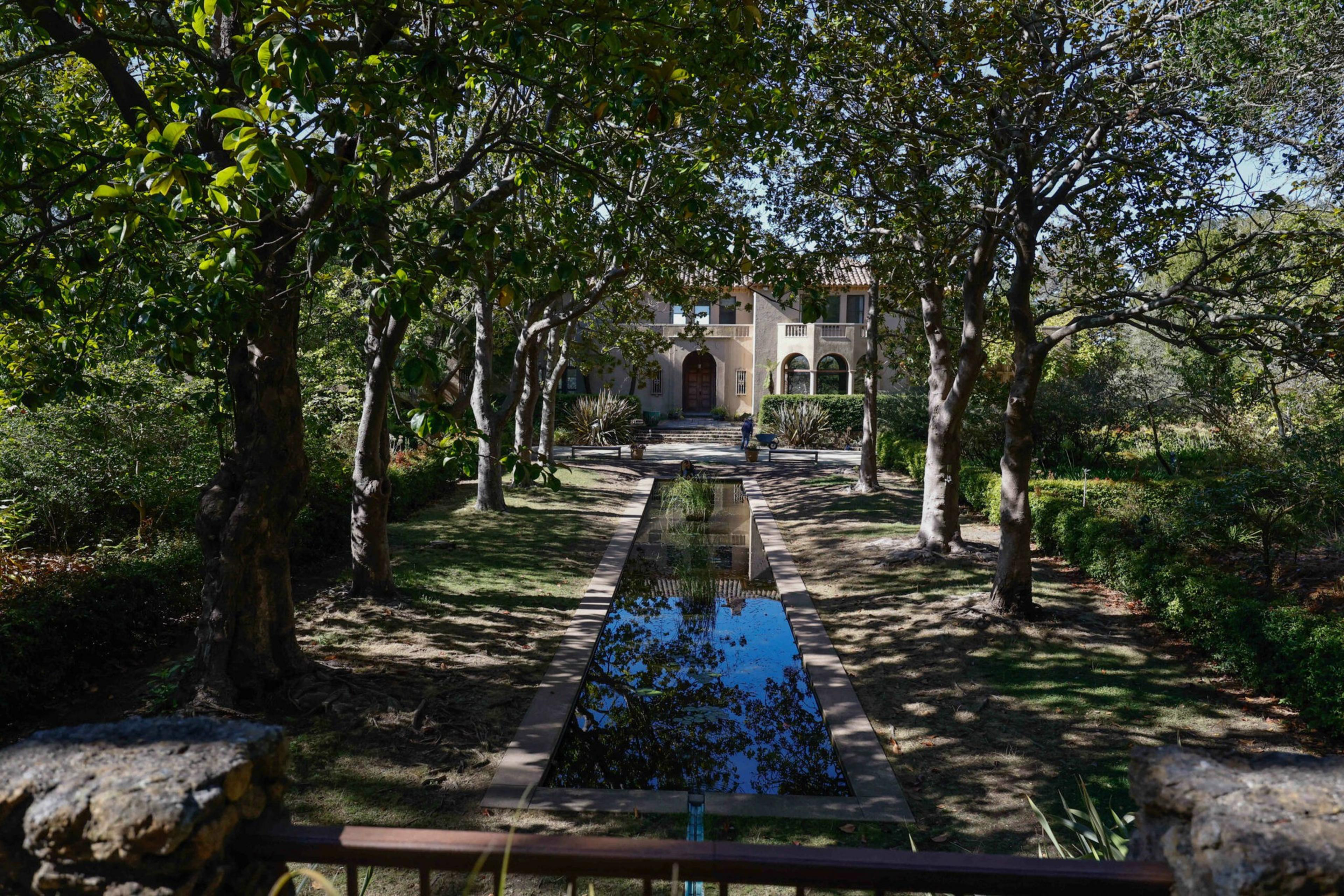 A stone building with arched windows and a wooden door sits at the end of a tree-lined path featuring a long reflecting pool.