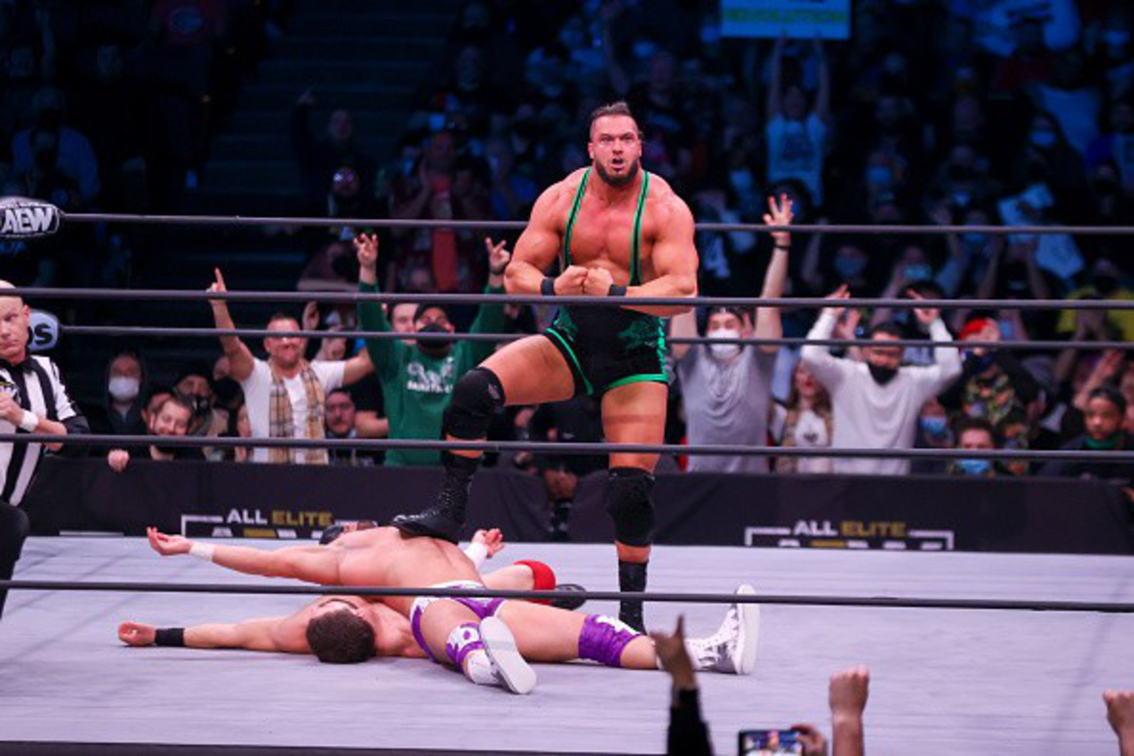 A muscular wrestler in green trunks stands victoriously with one foot on a defeated opponent lying face-up in the wrestling ring, with a cheering crowd in the background.