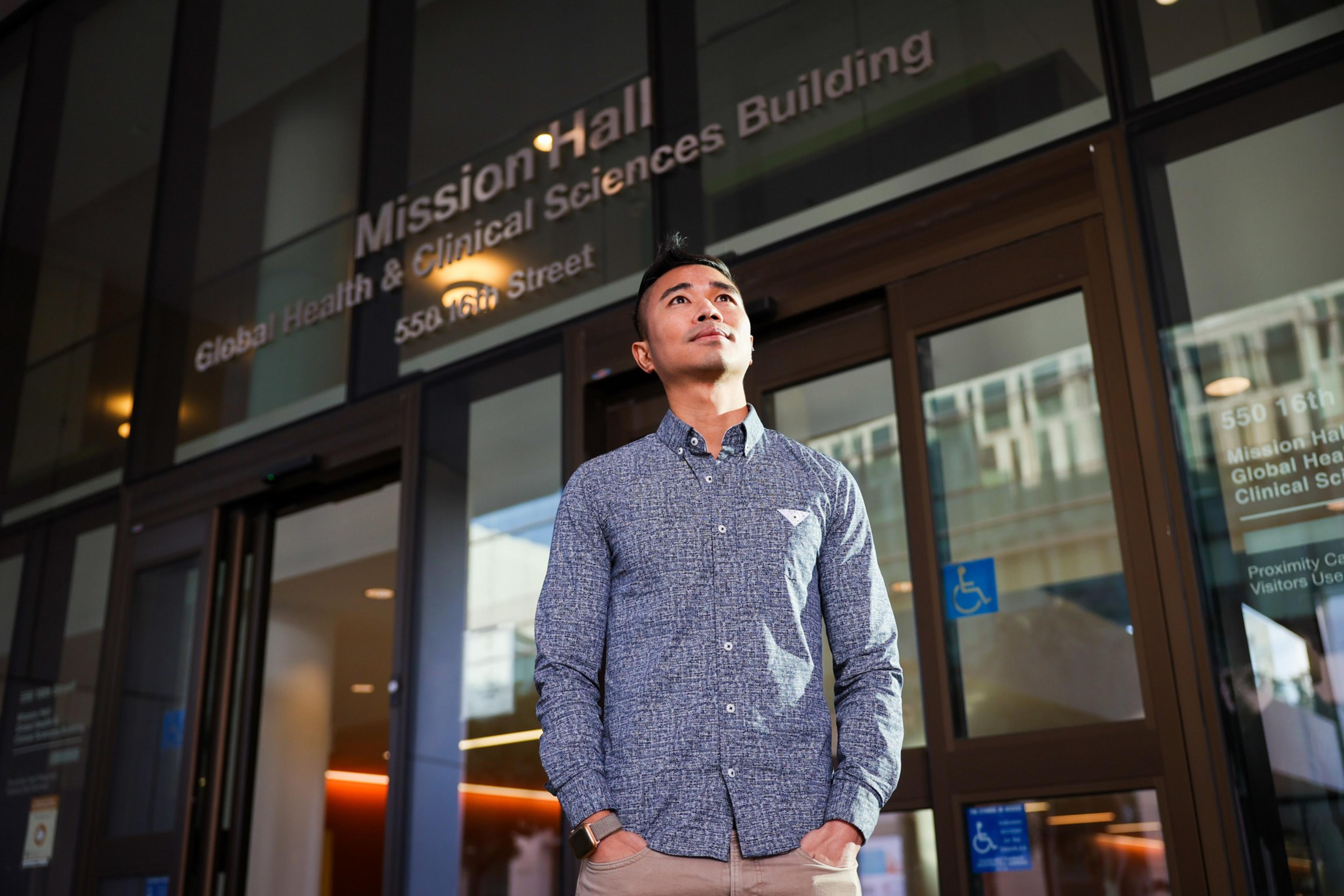 A man in a blue patterned shirt stands with hands in pockets, looking upward outside the Mission Hall Global Health & Clinical Sciences Building.