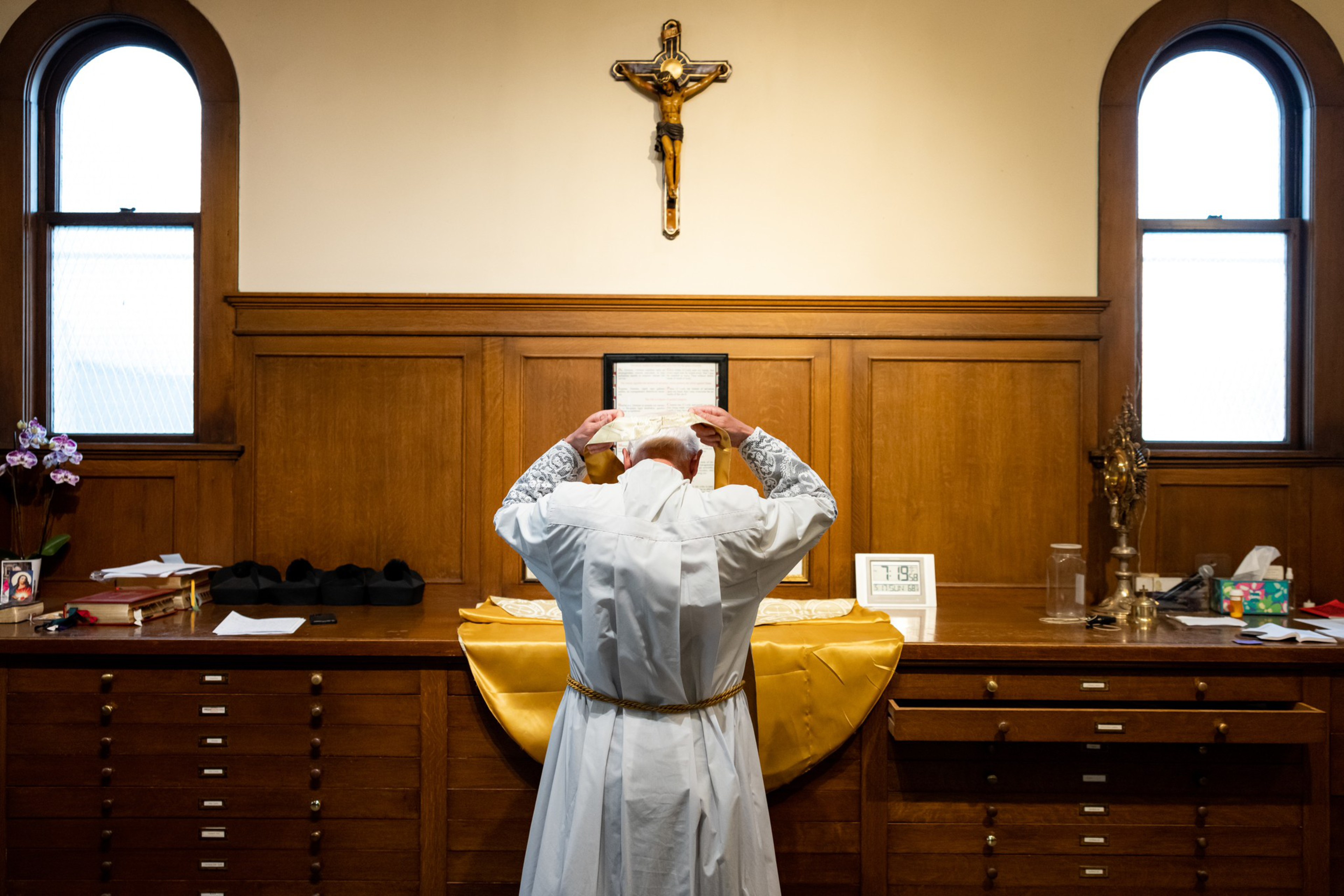 A priest dressed in white robes adjusts his headwear while standing before a wooden altar with religious items and a crucifix on the wall.