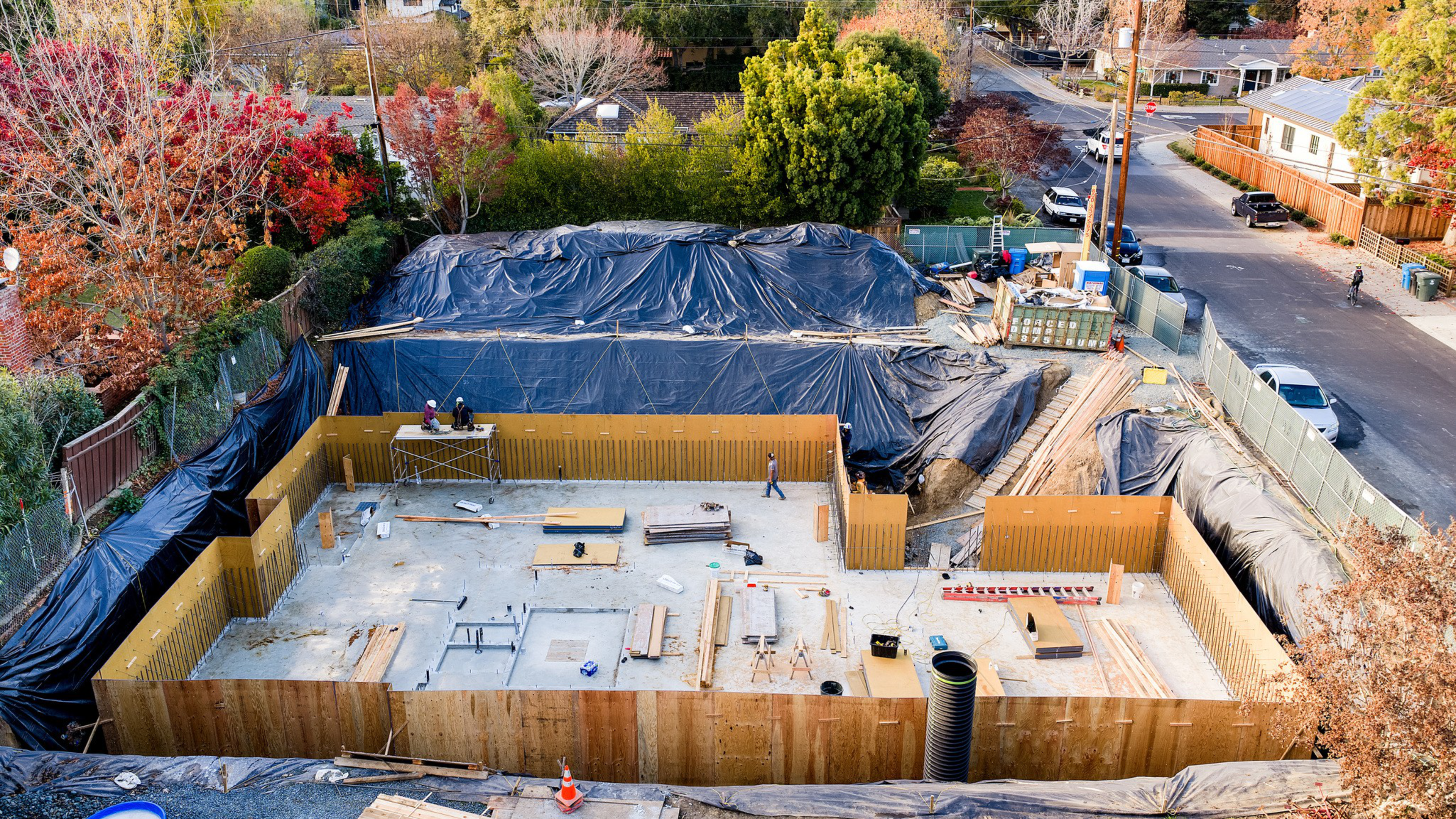 A large, rectangular construction site with wooden formwork and concrete slab, surrounded by trees, houses, and a street with parked cars.