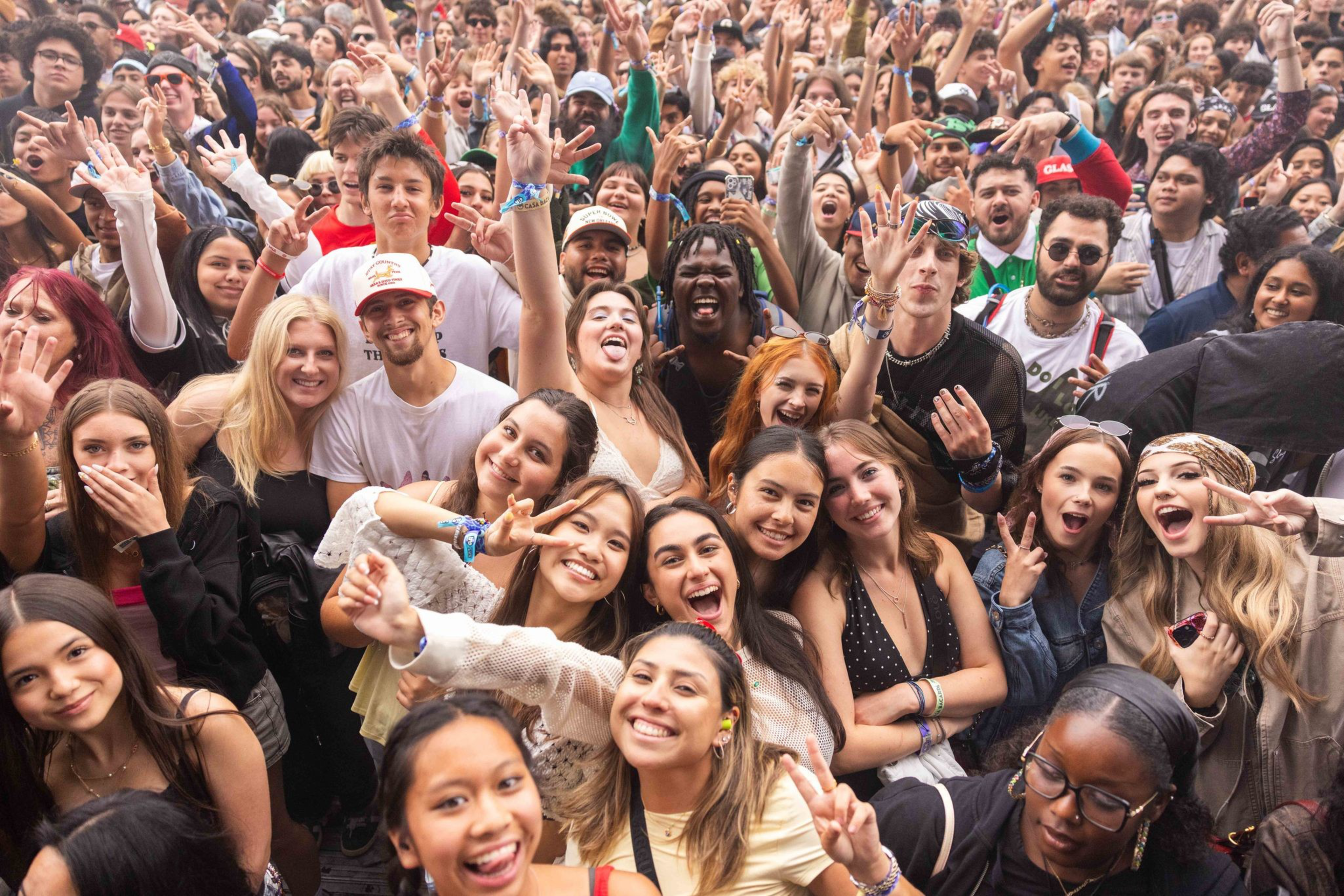 A large, diverse crowd of smiling young people cheer, wave, and make peace signs at a lively outdoor event or concert.