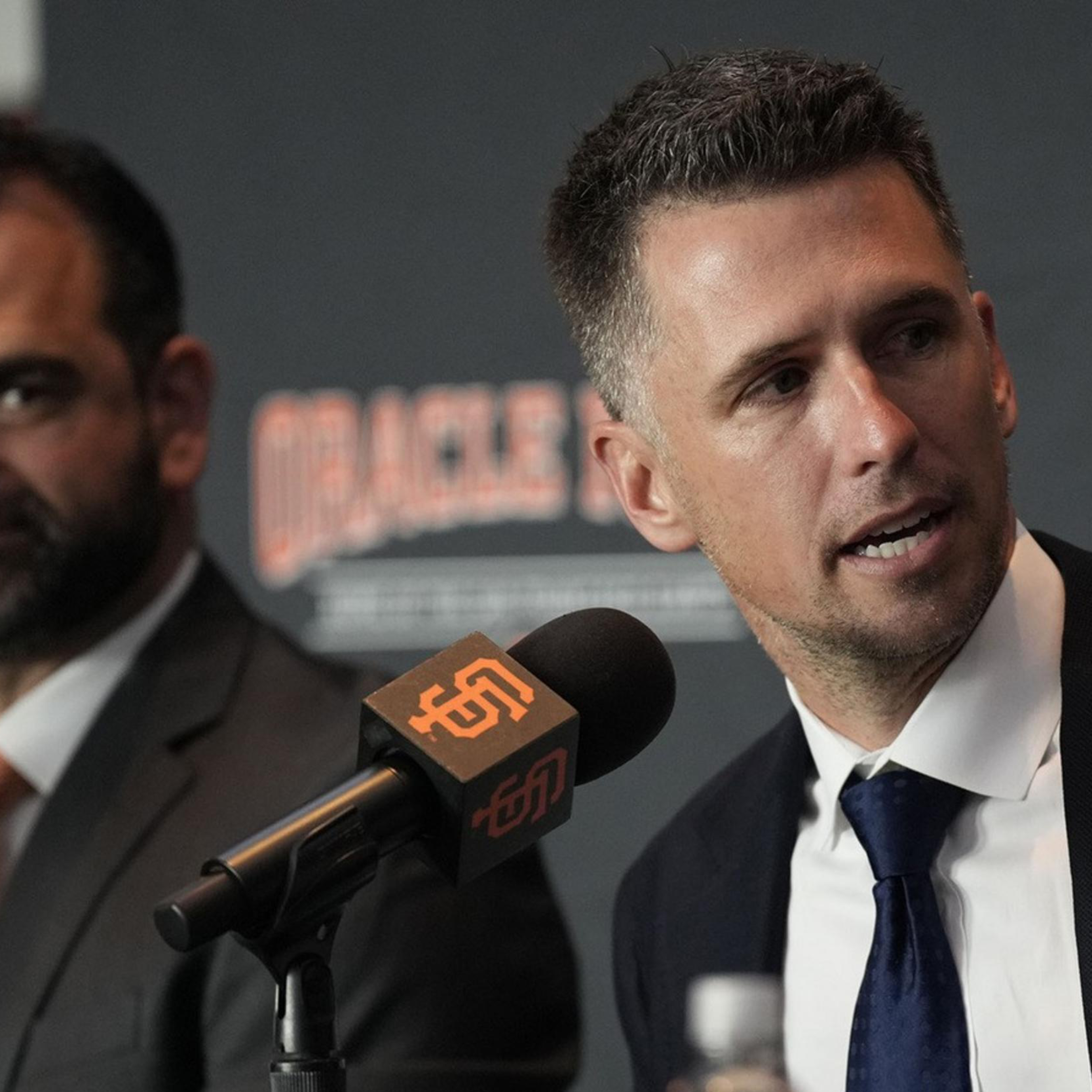 Two men in suits sit at a table with microphones labeled "Giants" in front of them. The man on the right is speaking while gesturing with his hand.
