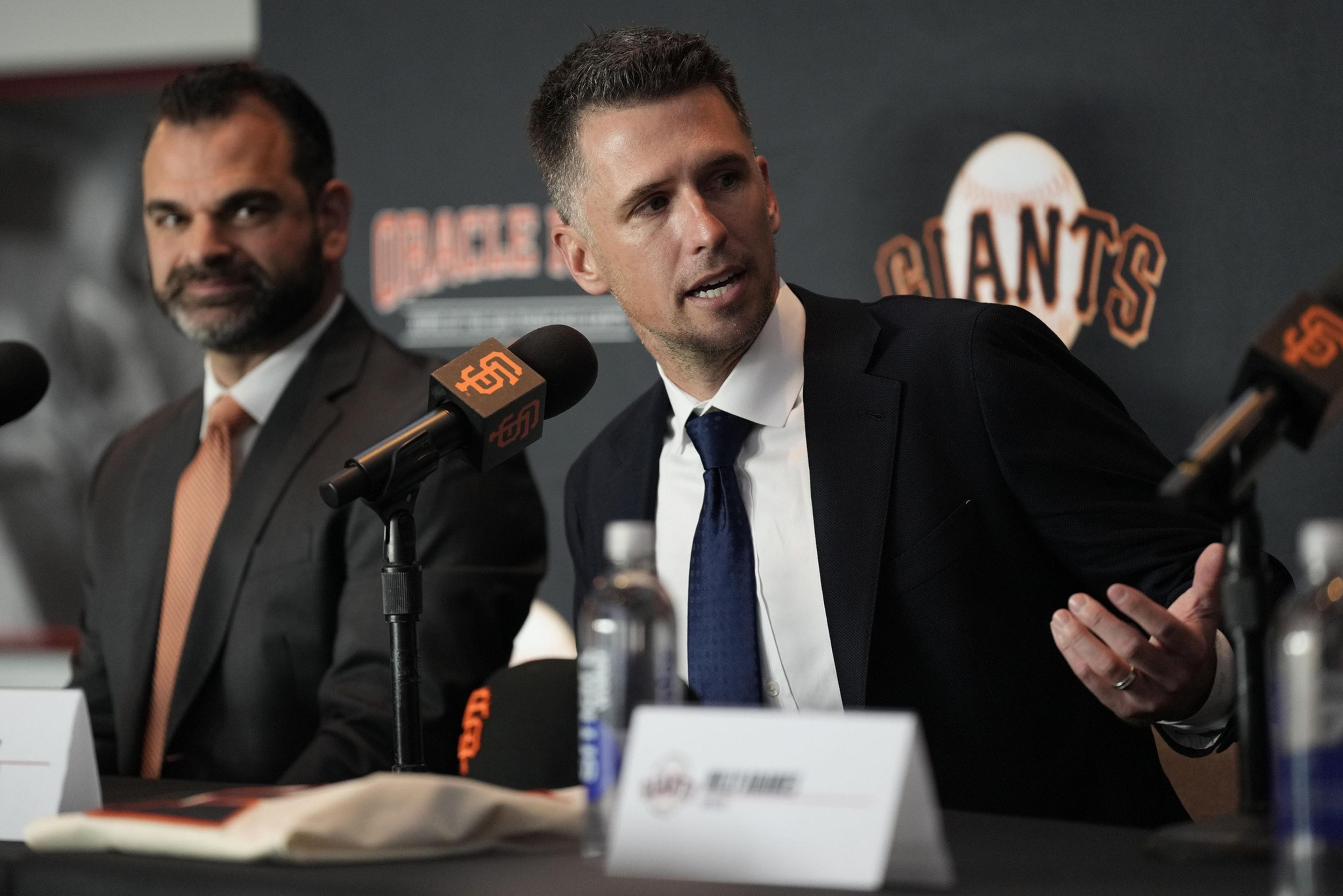 Two men in suits sit at a table with microphones labeled "Giants" in front of them. The man on the right is speaking while gesturing with his hand.