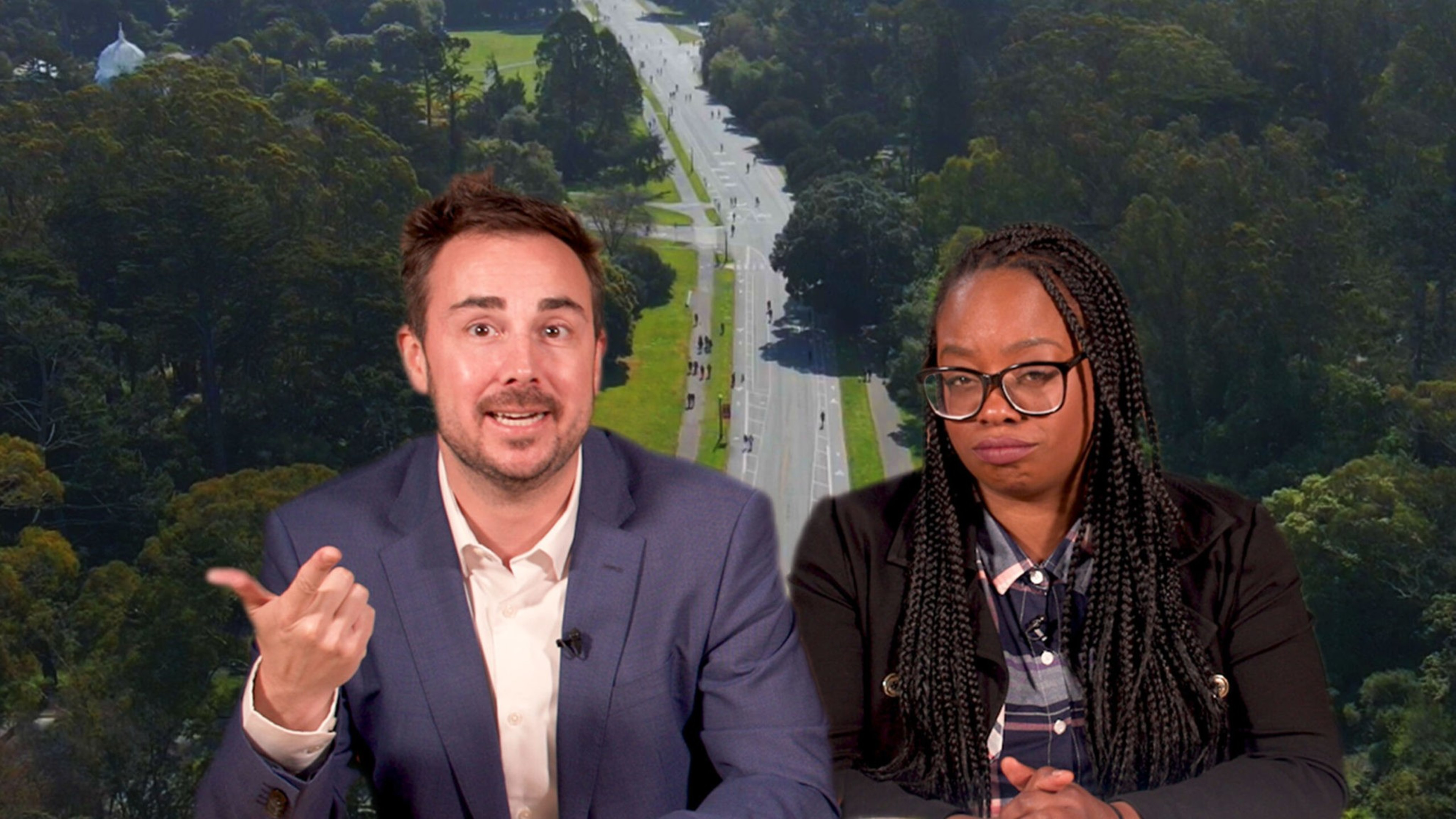 A man in a blue suit gestures while speaking, and a woman with glasses and braided hair sits beside him with a serious expression.