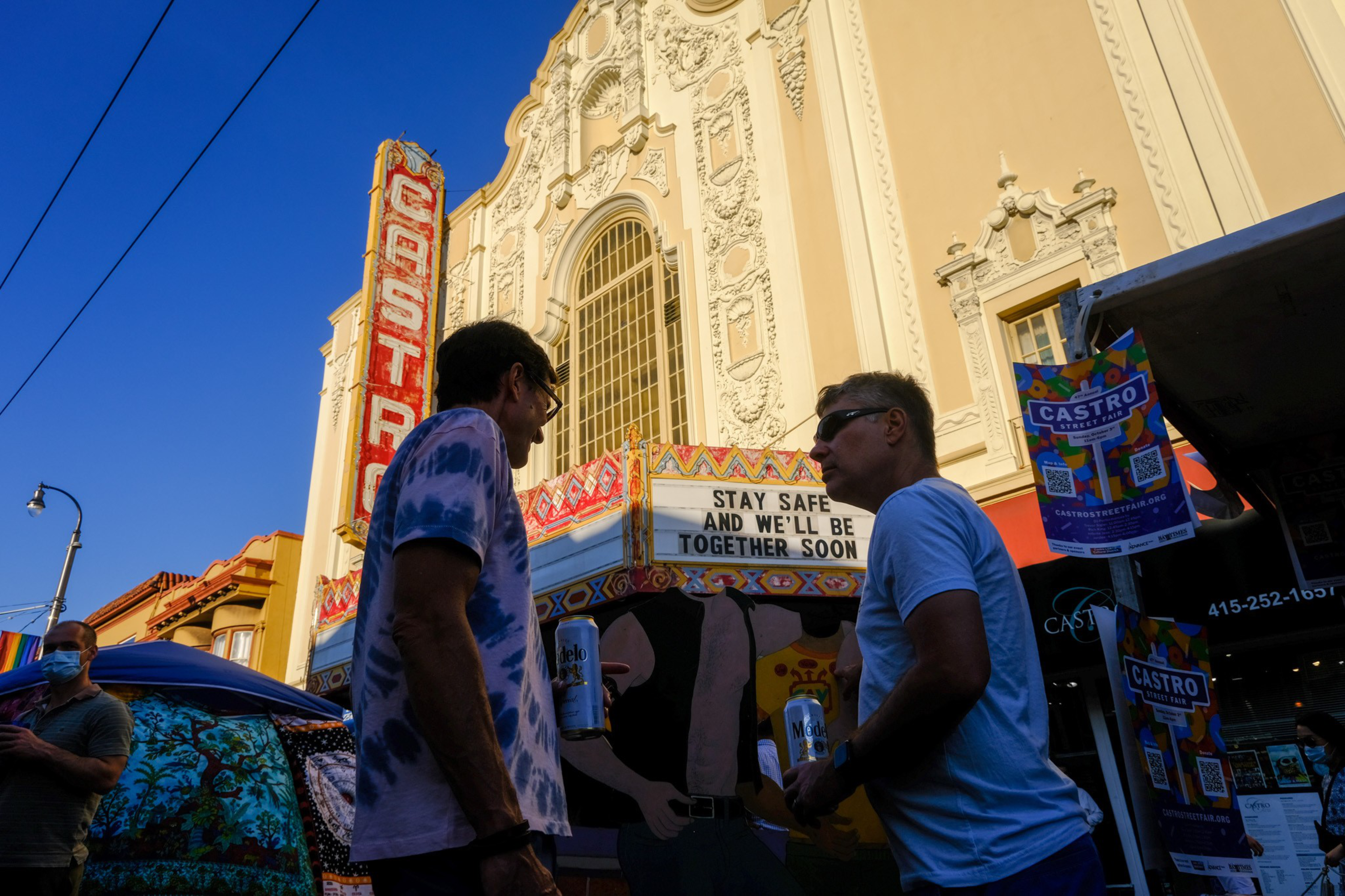 Two men hold drinks and chat outside the ornate Castro Theatre, with a marquee reading "Stay Safe and We'll Be Together Soon."