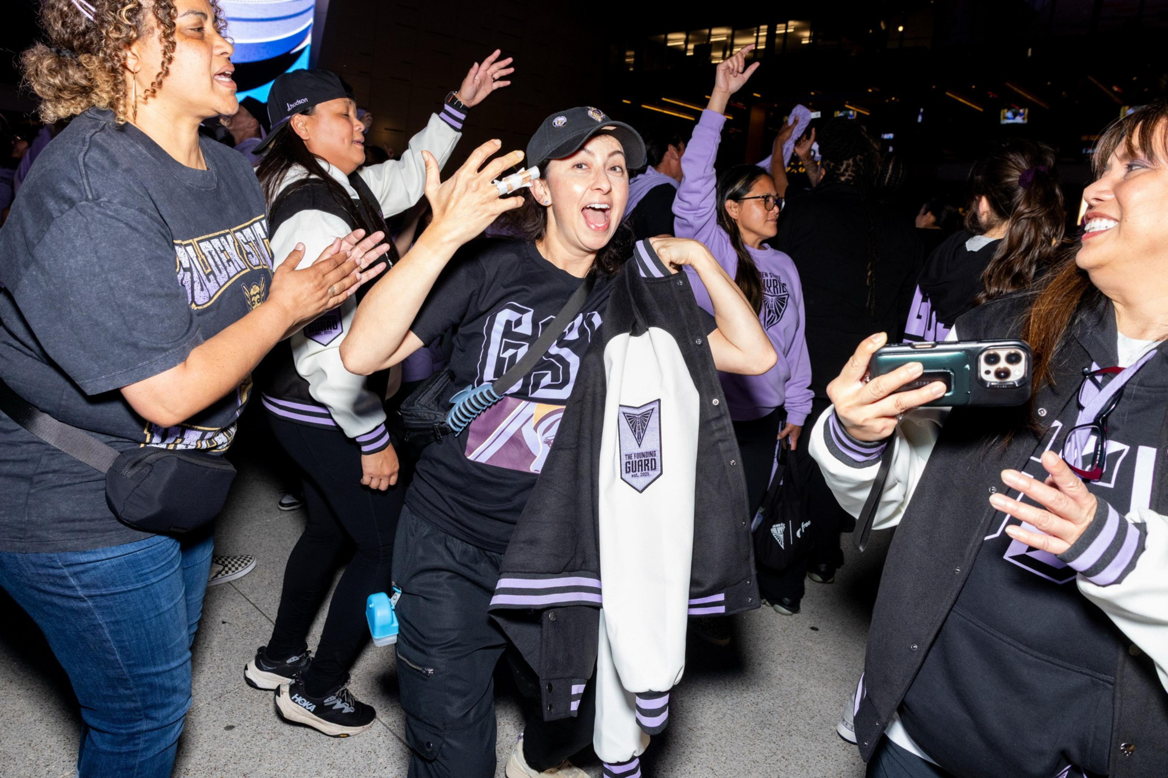 A group of excited people wearing purple and black sports gear cheer and take photos, holding up a jacket with a "The Guard" logo.