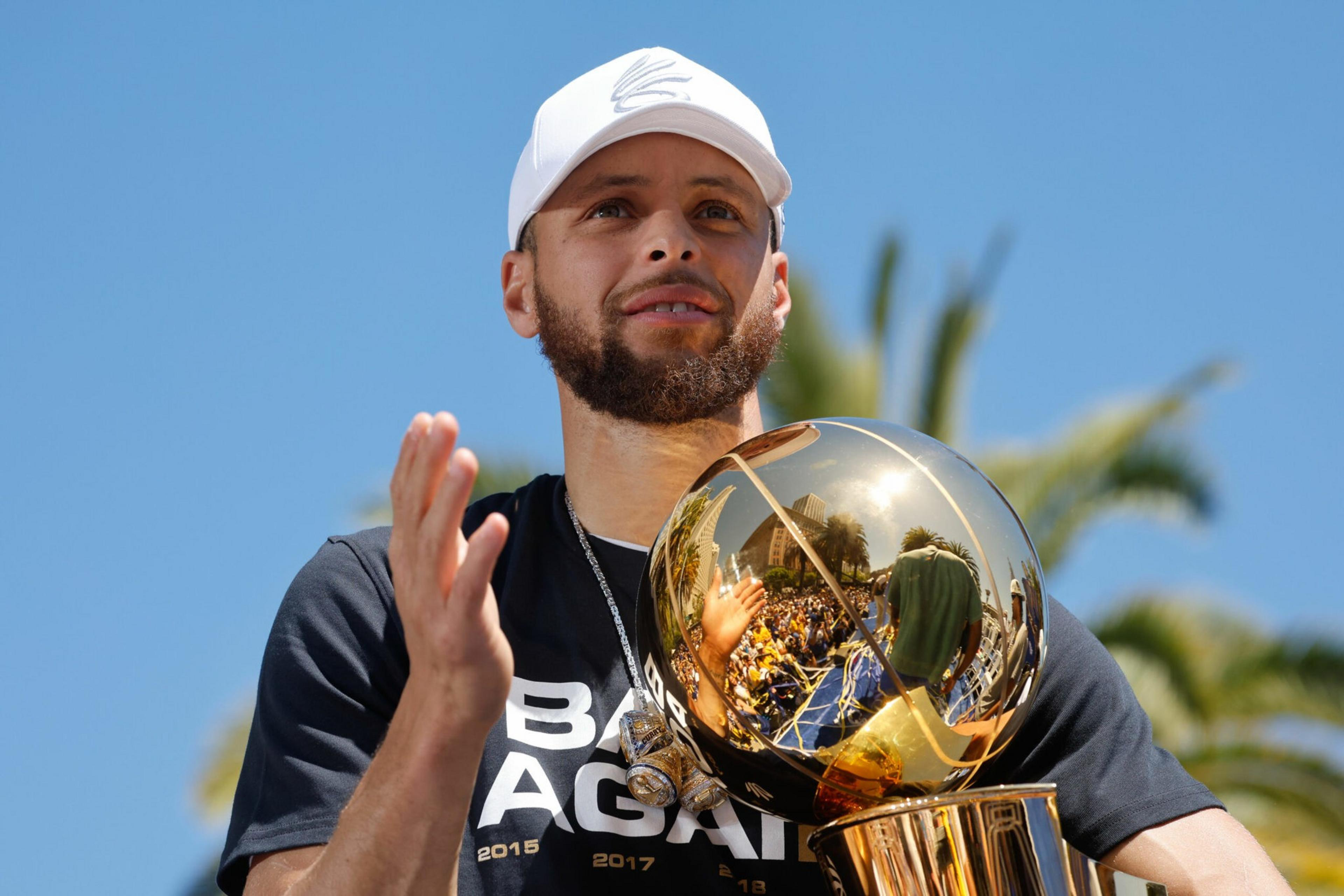 A man with a beard in a white cap and black shirt claps while holding a shiny gold basketball trophy outdoors under a clear blue sky.
