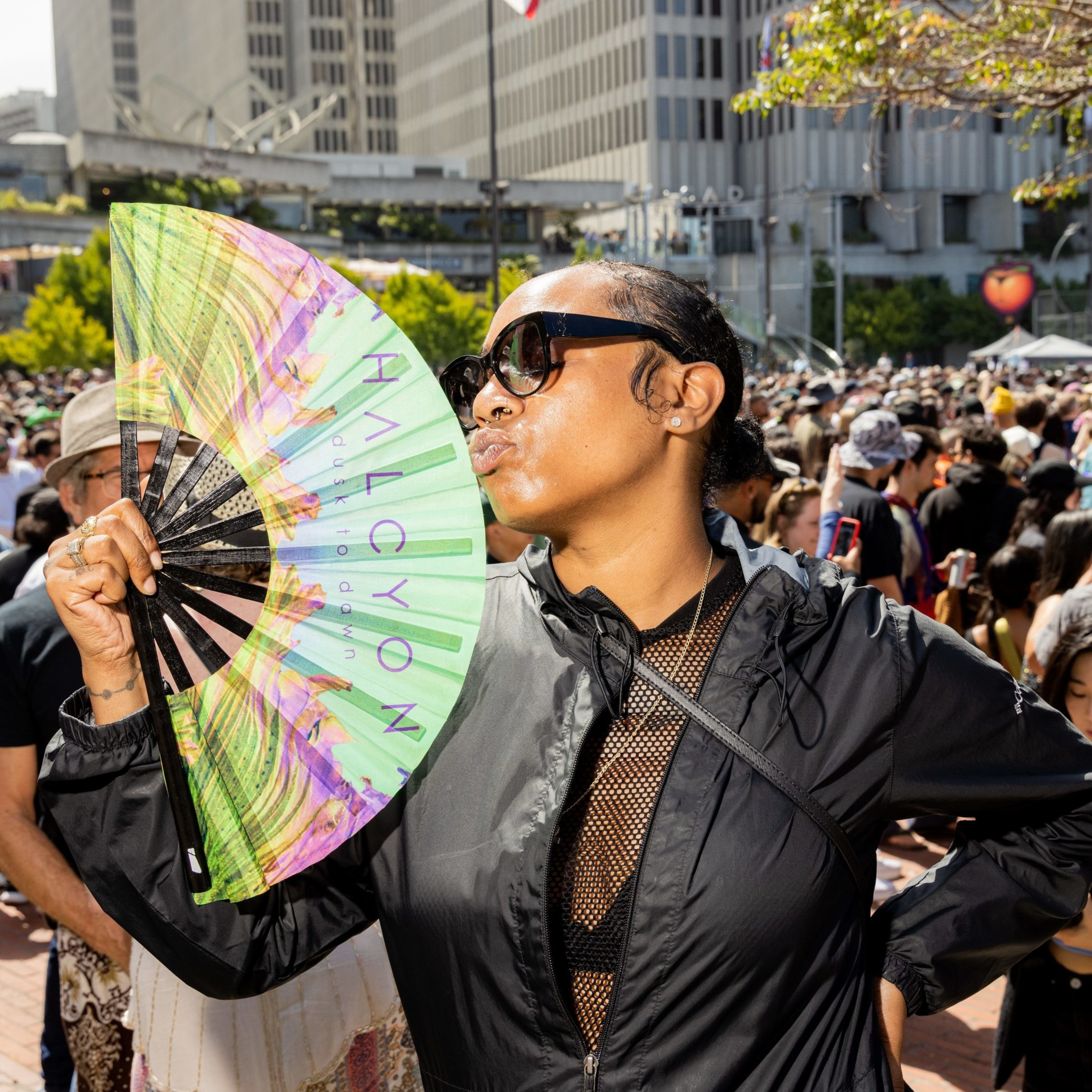 A person in sunglasses and a black jacket is holding a colorful fan and making a playful facial expression in front of a large, outdoor crowd in a city setting.