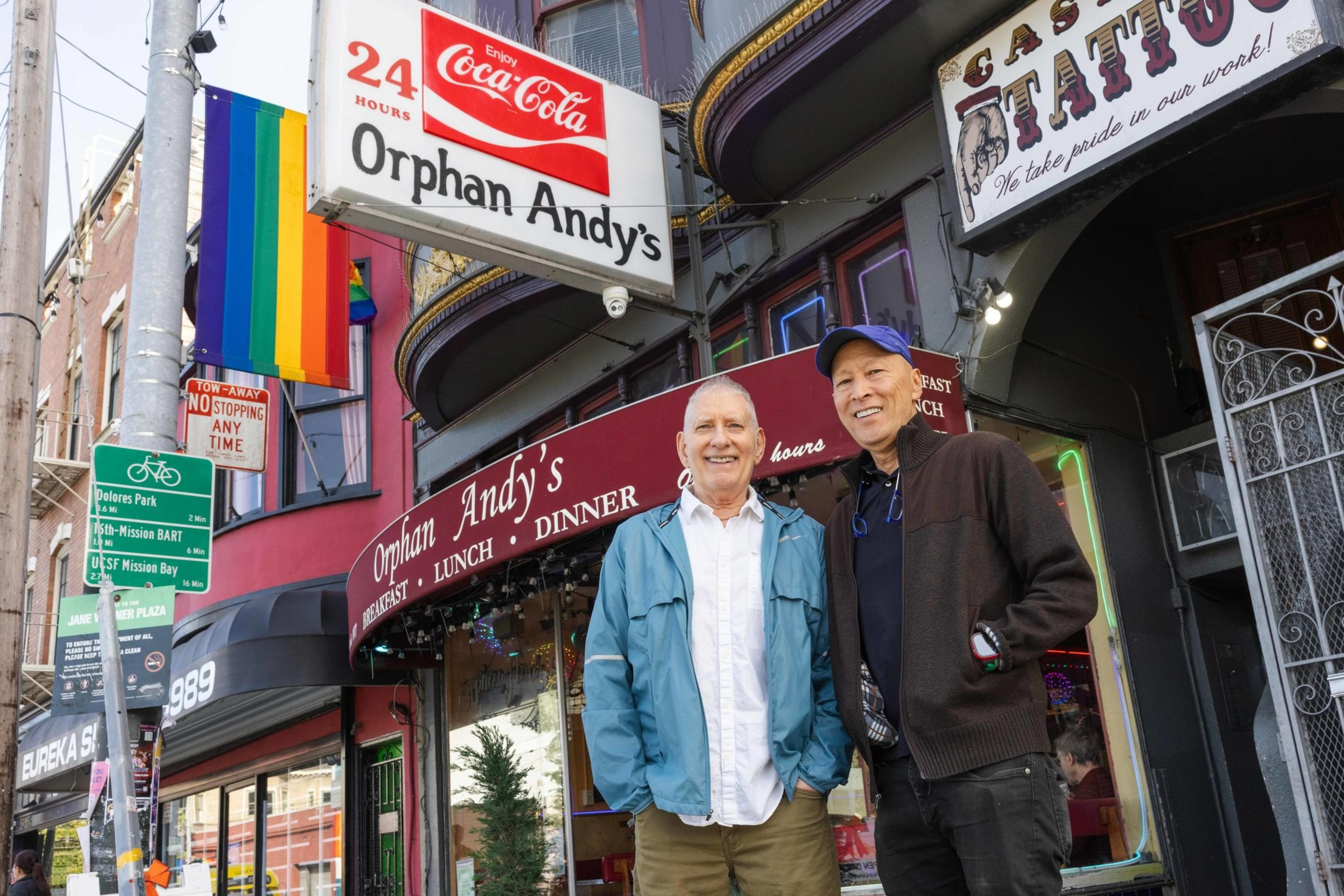 Two people are smiling in front of Orphan Andy's, a diner with a Coca-Cola sign and a rainbow flag. The street is vibrant with various signs and colors.