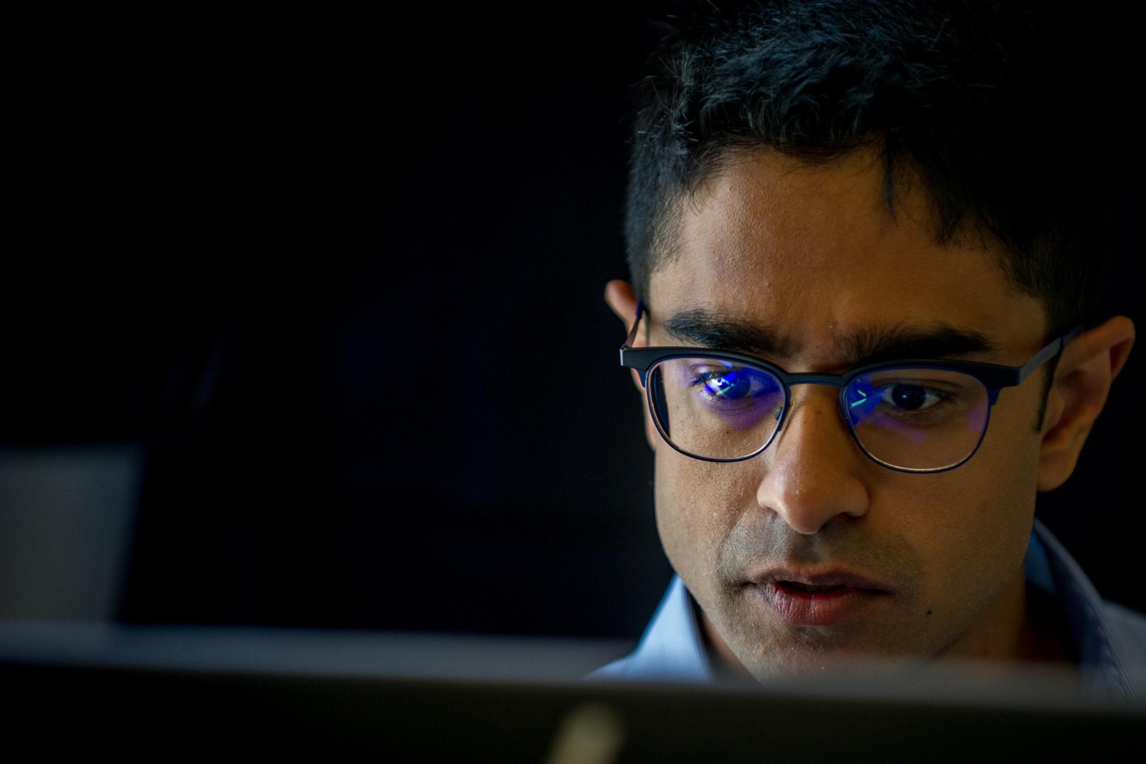 A person with glasses is intensely focused on something, likely a computer screen, which is reflected in the glasses. The background is dark and blurred.
