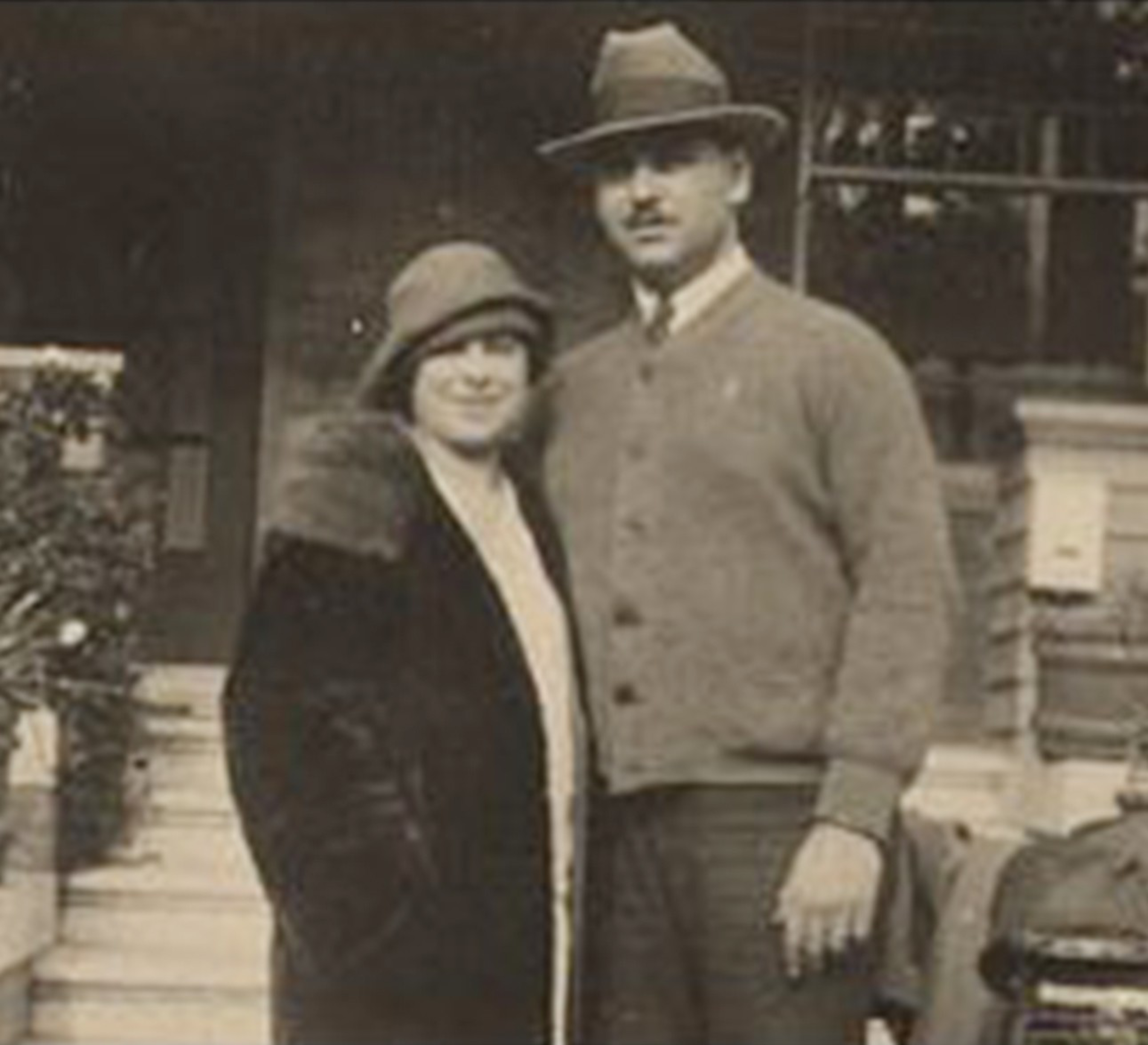 A man and woman stand close together outside a building; the woman wears a cloche hat and fur-collared coat, the man wears a fedora and buttoned cardigan.