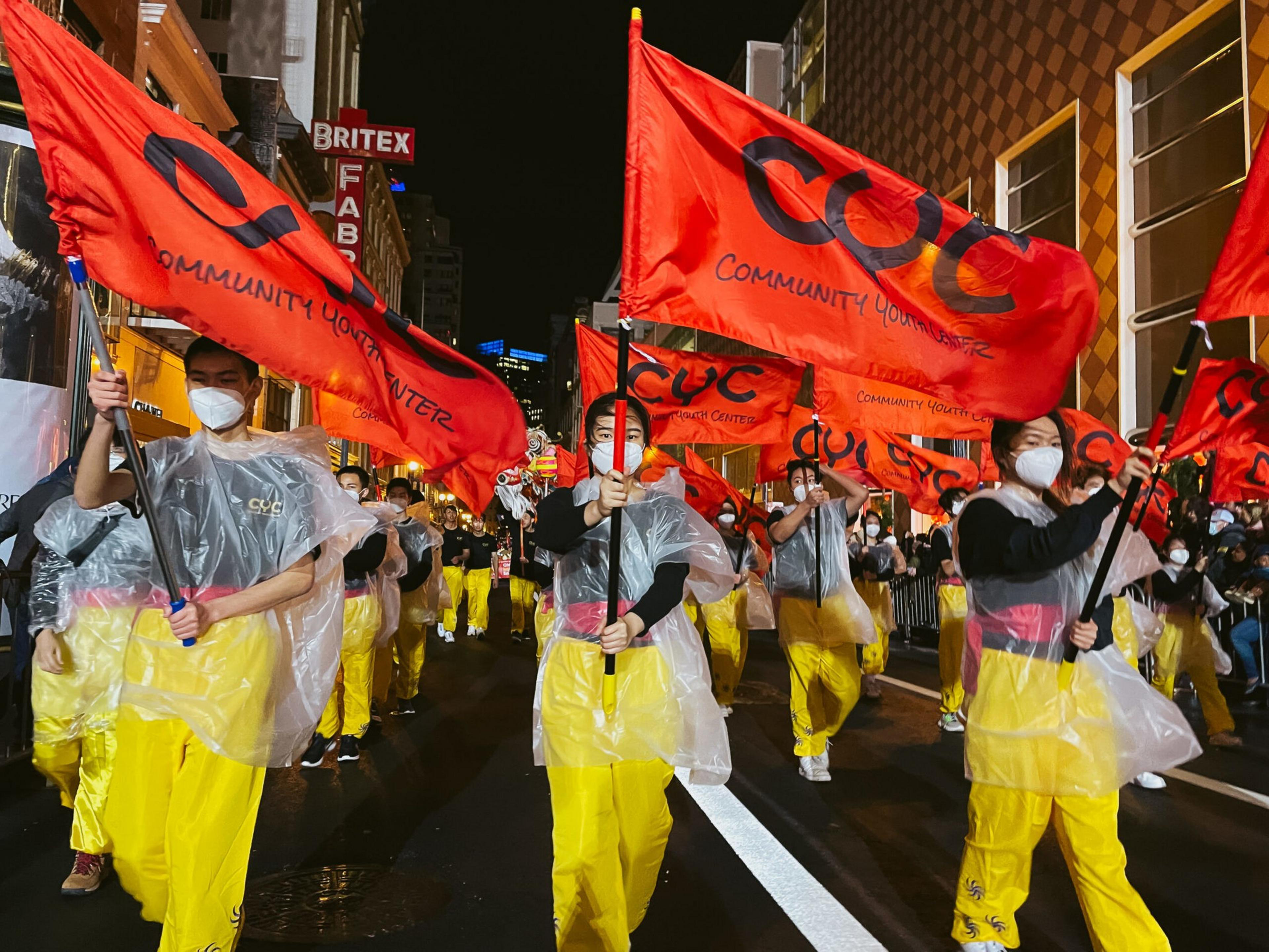 A group of people wearing yellow pants, black shirts, white masks, and clear ponchos march at night holding large red flags reading "Community Youth Center."