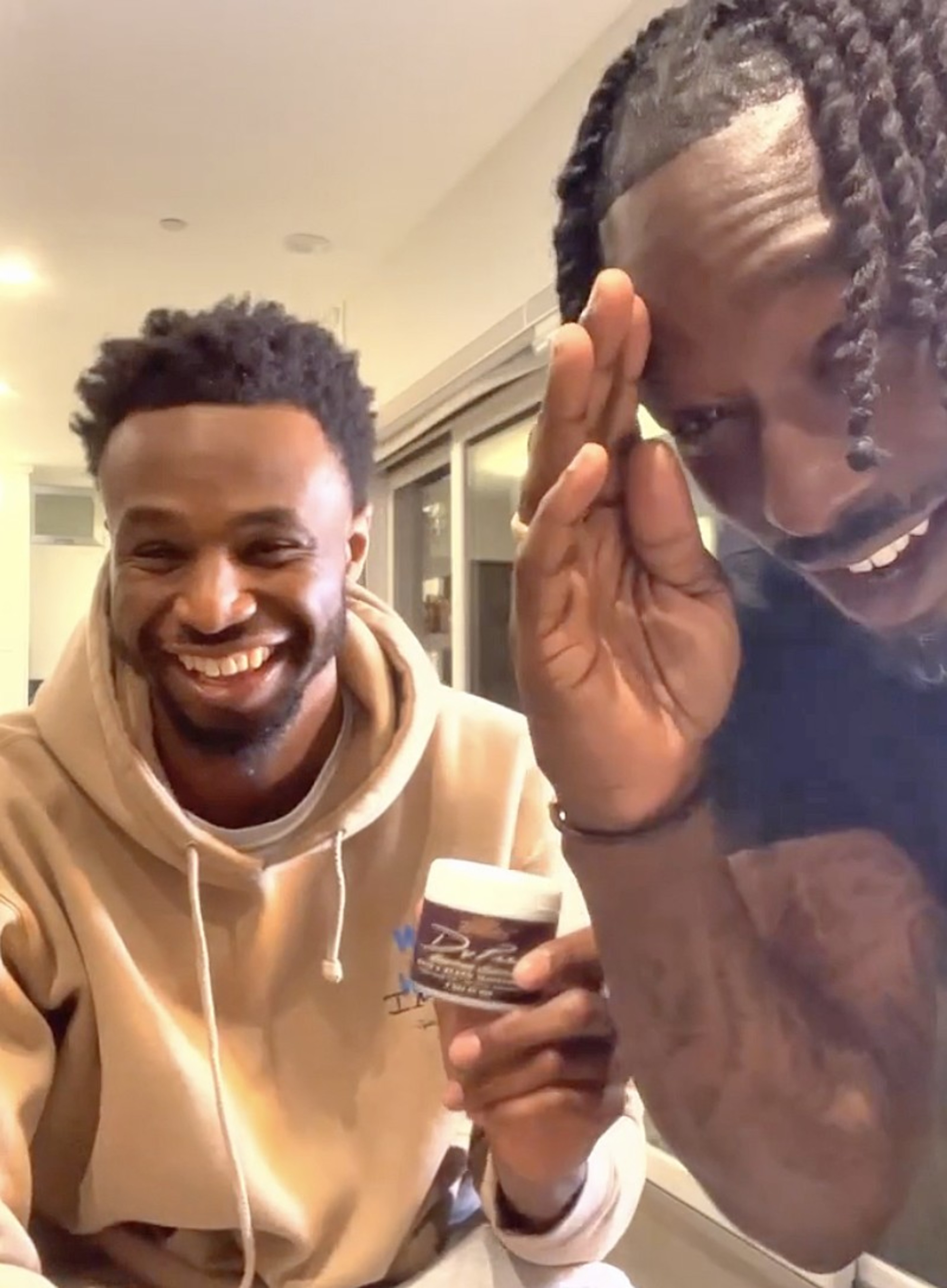 Two men are smiling and gesturing, one holding a product. They're indoors, sharing a light-hearted moment.