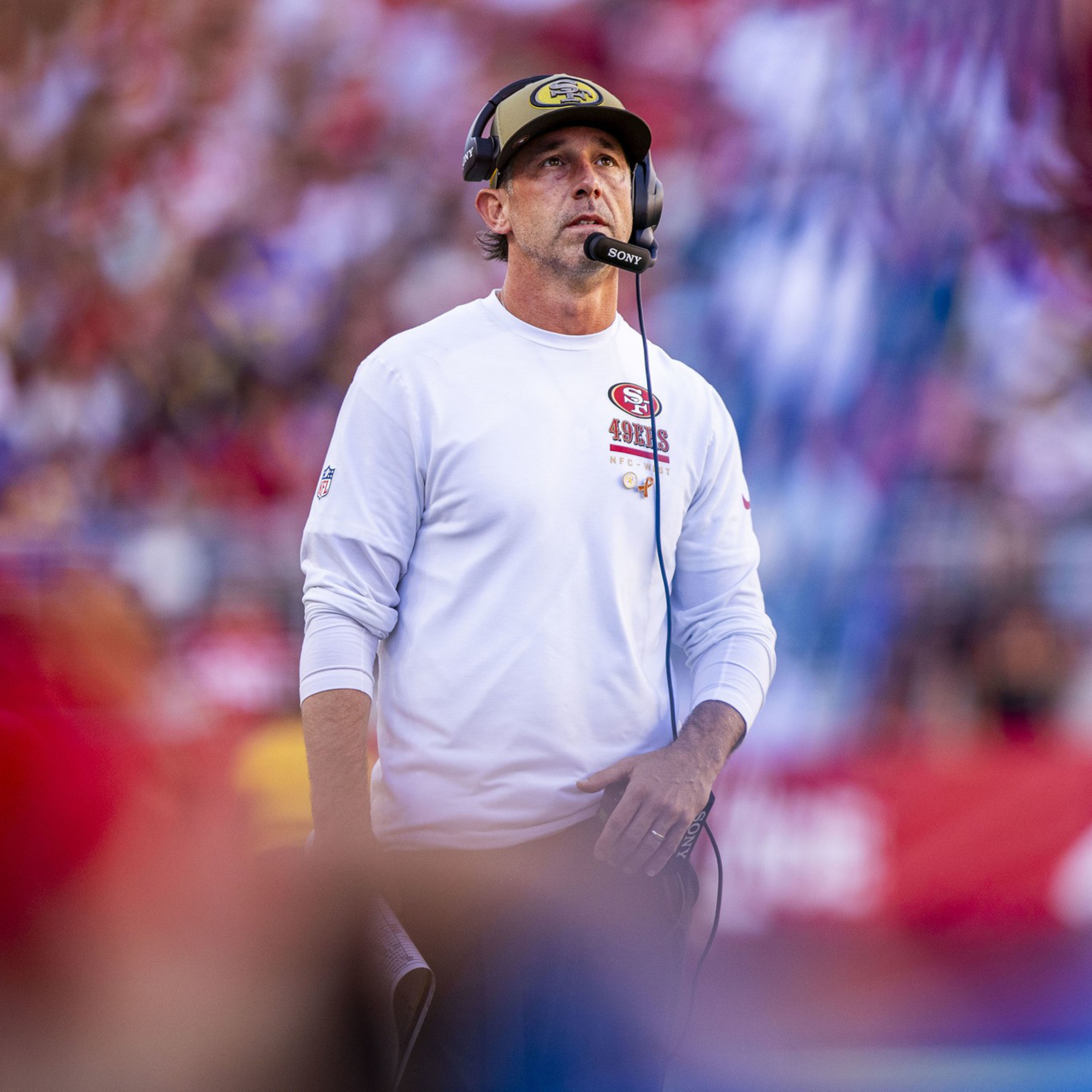 A man wearing a white 49ers long-sleeve shirt, headset, and a cap stands on a blurred football field with fans in the background.