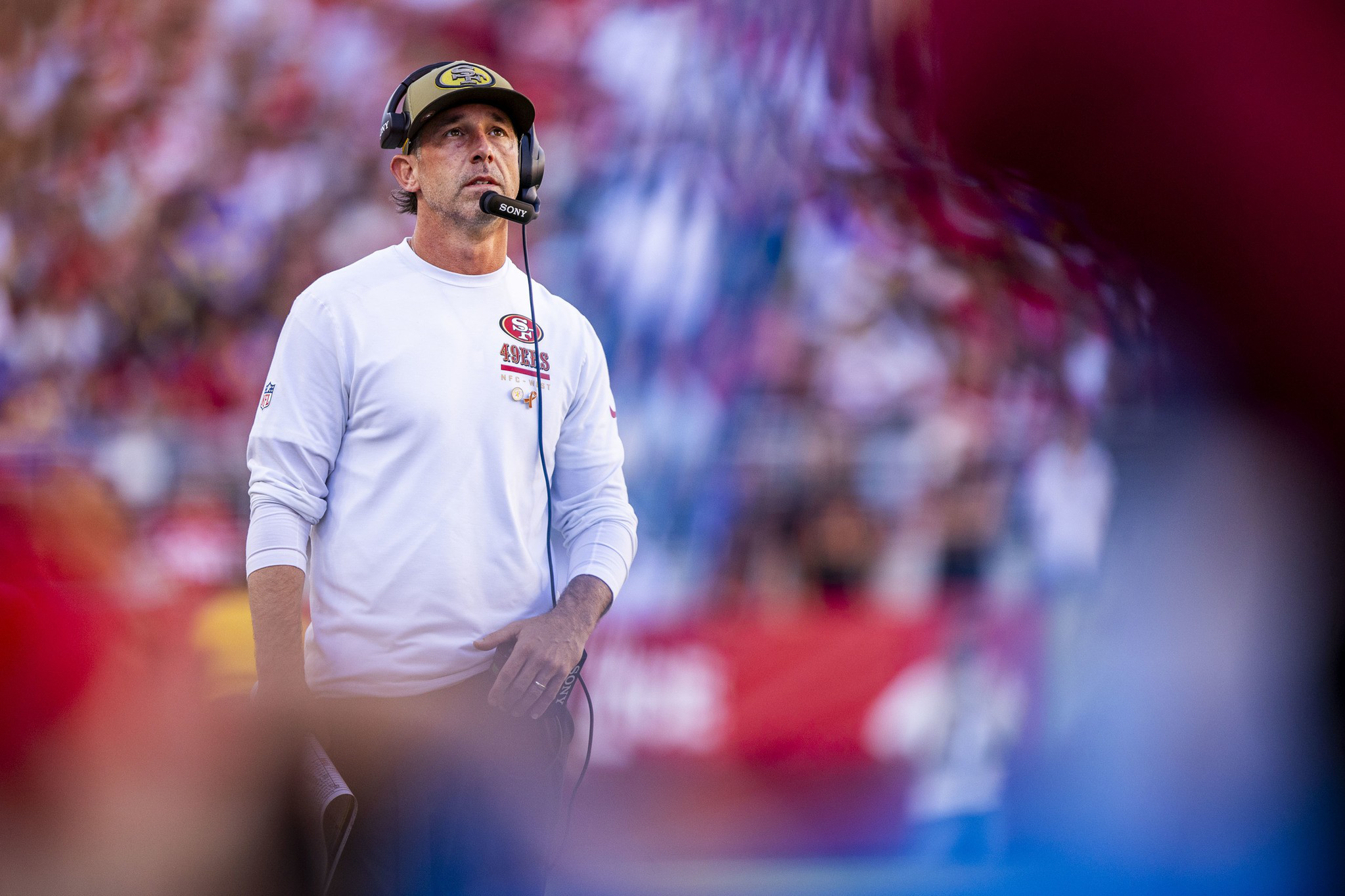 A man wearing a white 49ers long-sleeve shirt, headset, and a cap stands on a blurred football field with fans in the background.