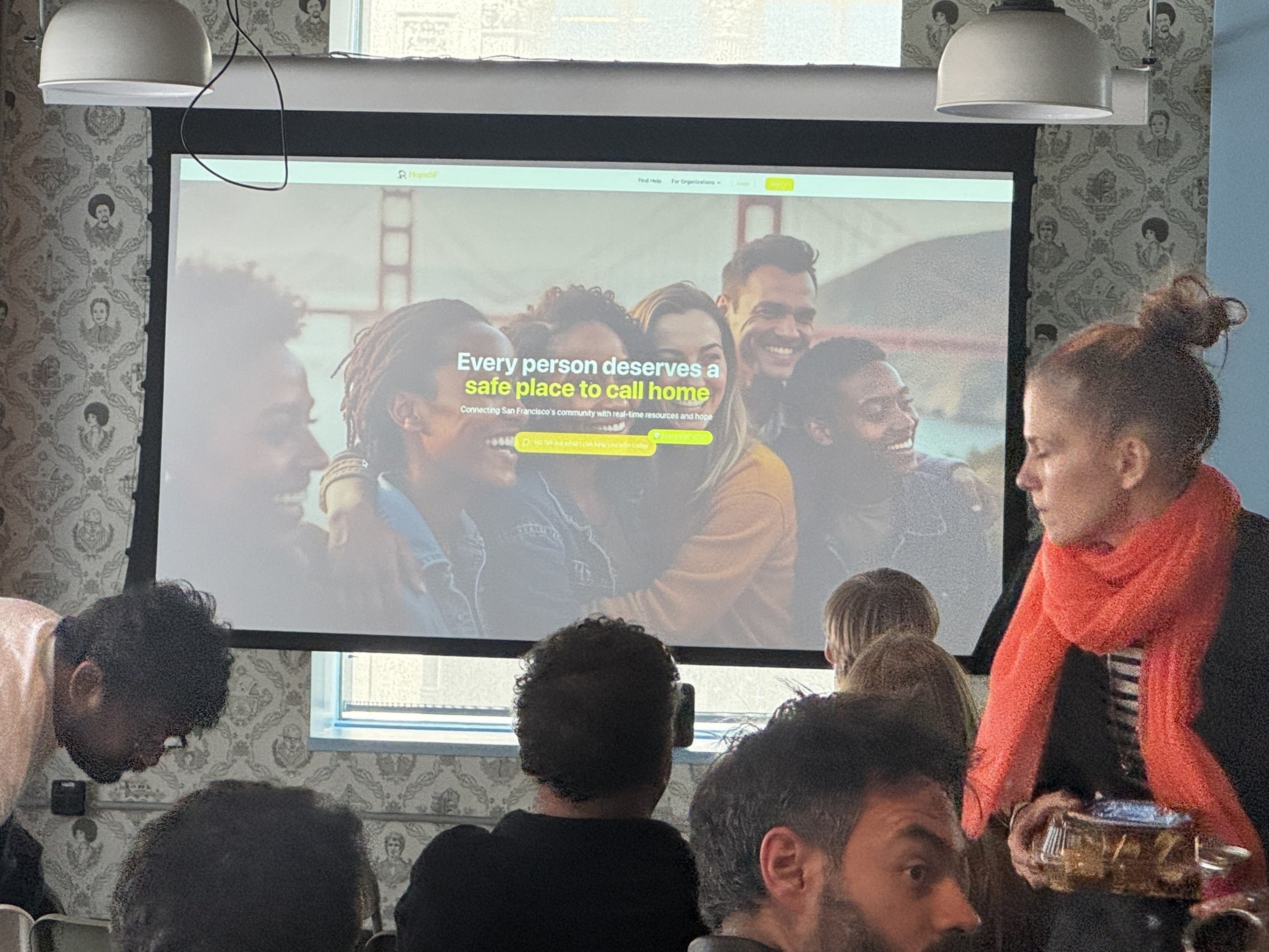A group of people faces a presentation screen with a webpage stating, "Every person deserves a safe place to call home," in a cozy room with patterned wallpaper.
