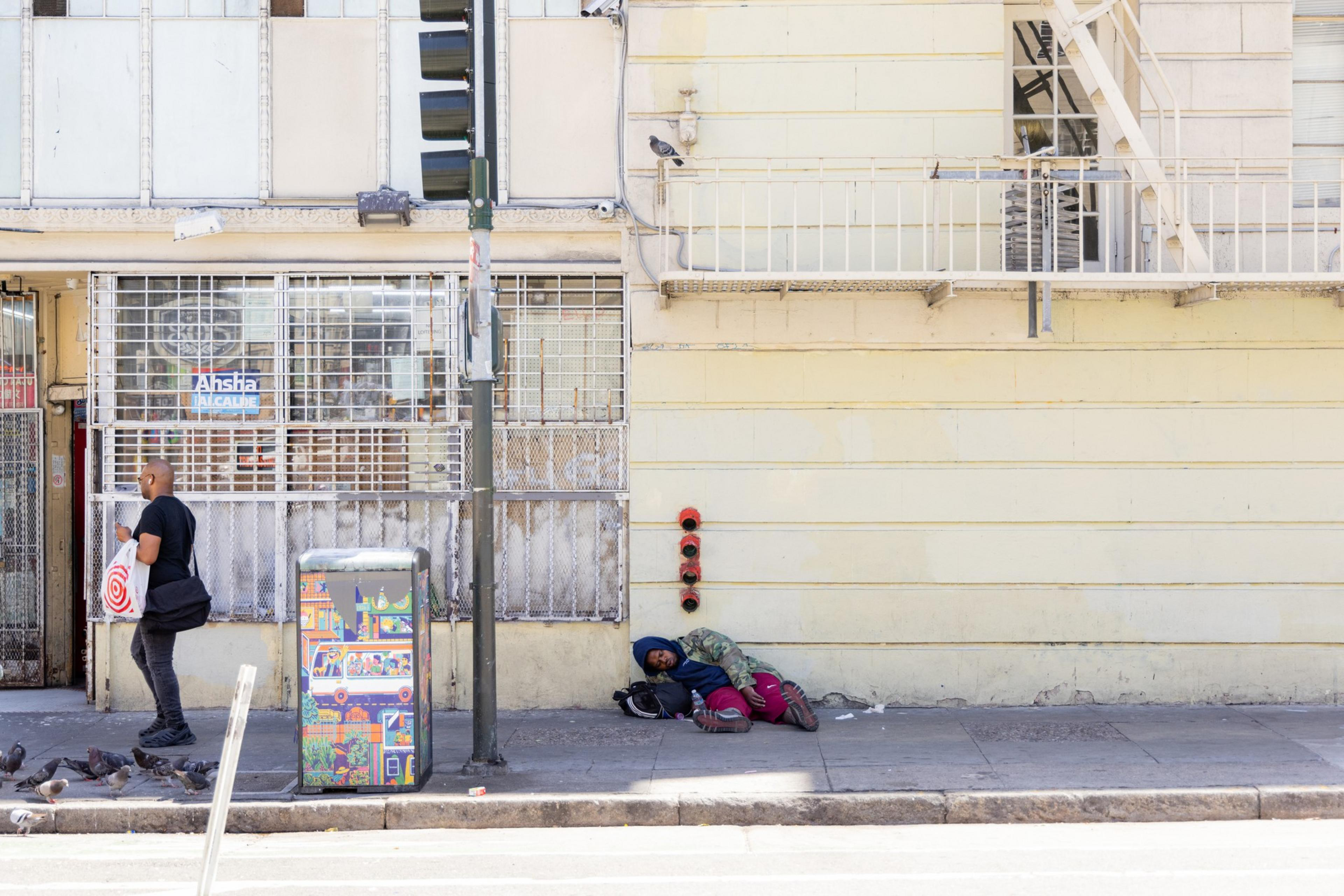 A person carrying a red-striped shopping bag walks past a sitting person under a beige building with barred windows. Birds peck at the ground nearby.