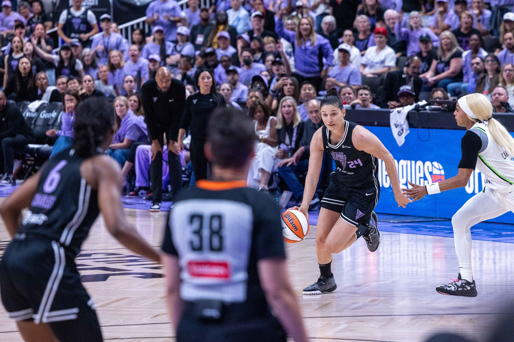 A basketball player in a black uniform dribbles the ball while facing a defender in a white uniform, with teammates, a referee, and a crowded audience in the background.