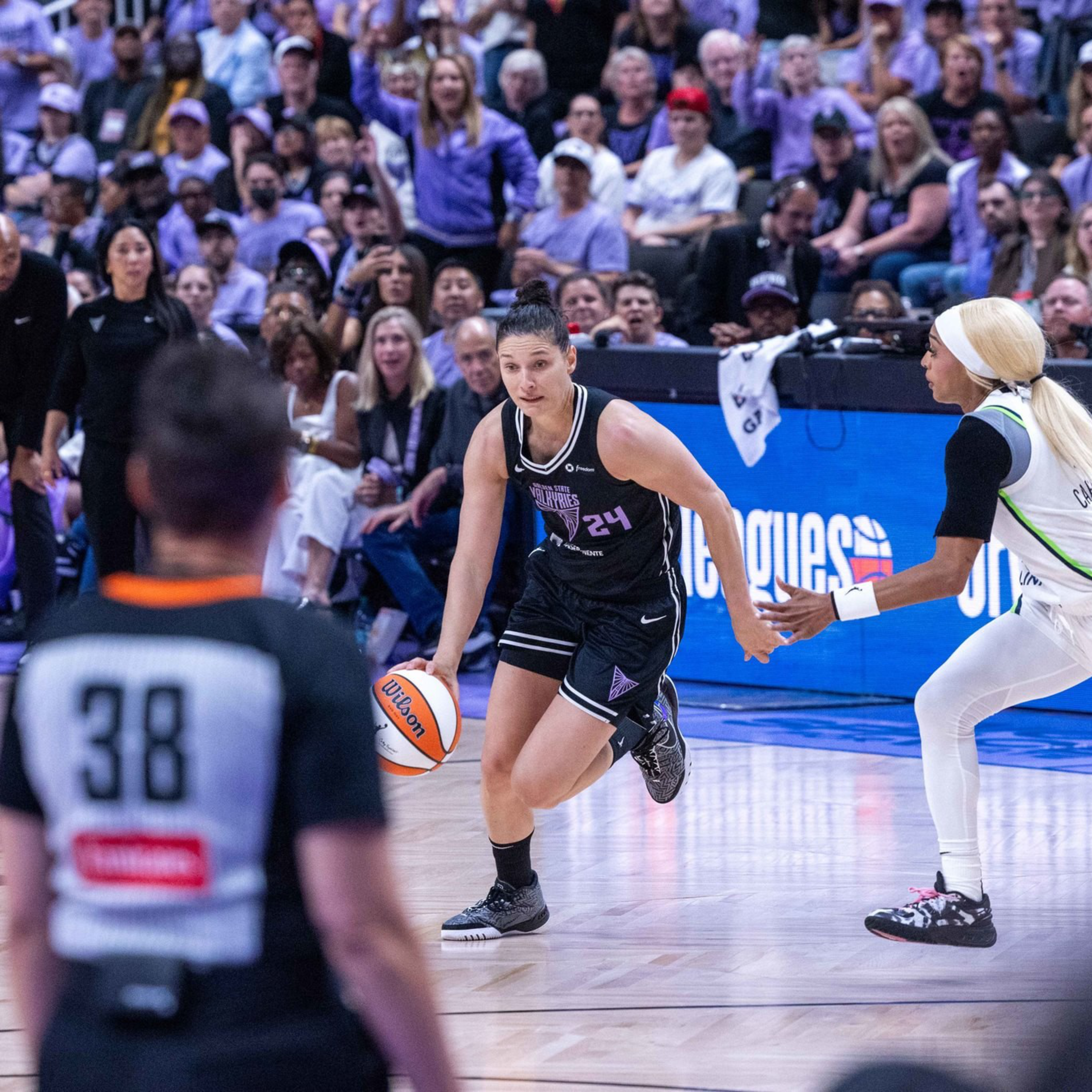 A basketball player in a black uniform dribbles the ball while facing a defender in a white uniform, with teammates, a referee, and a crowded audience in the background.