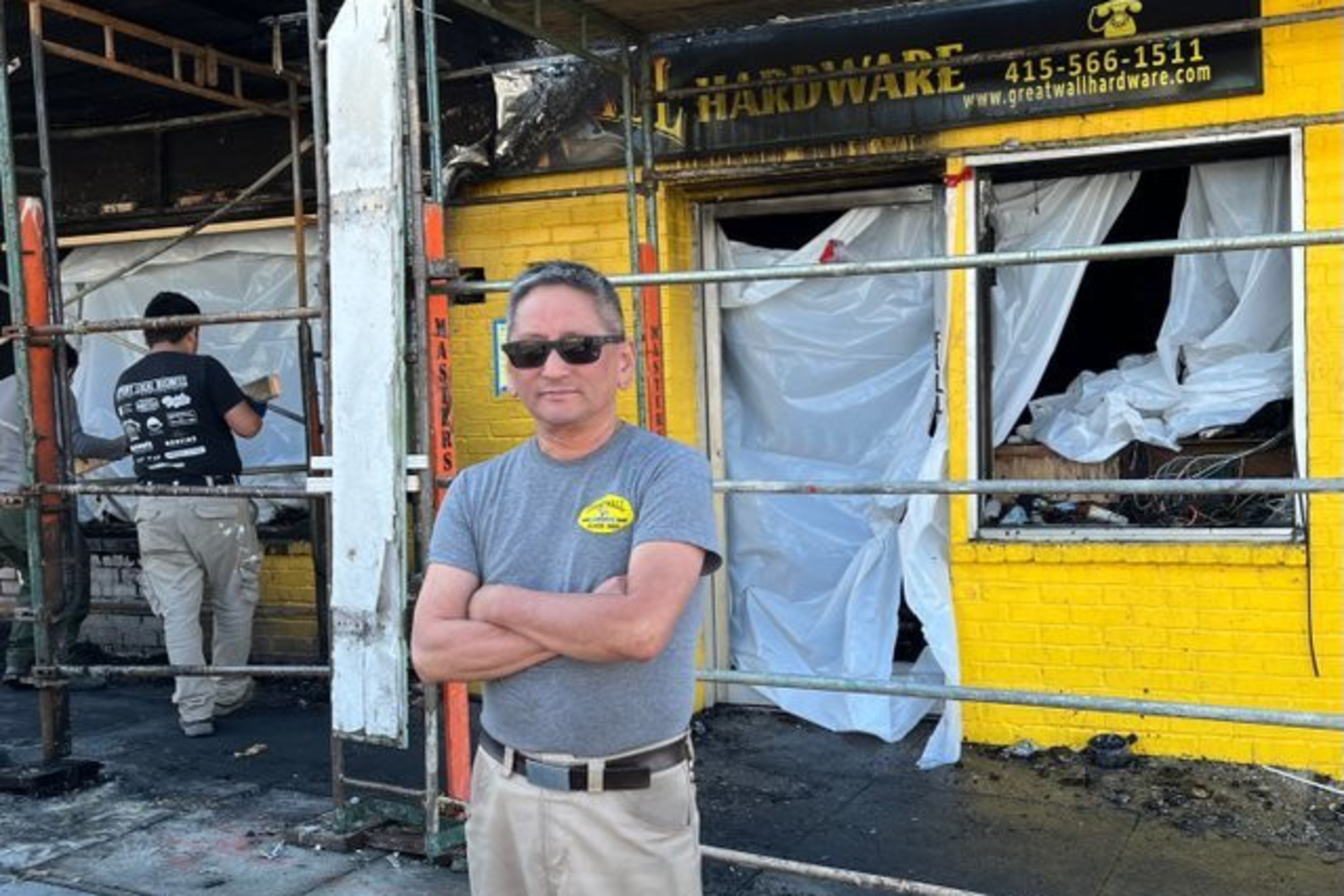 A man in sunglasses stands with arms crossed in front of a burnt yellow hardware store with plastic covering its windows. Another man is working on the damaged store.