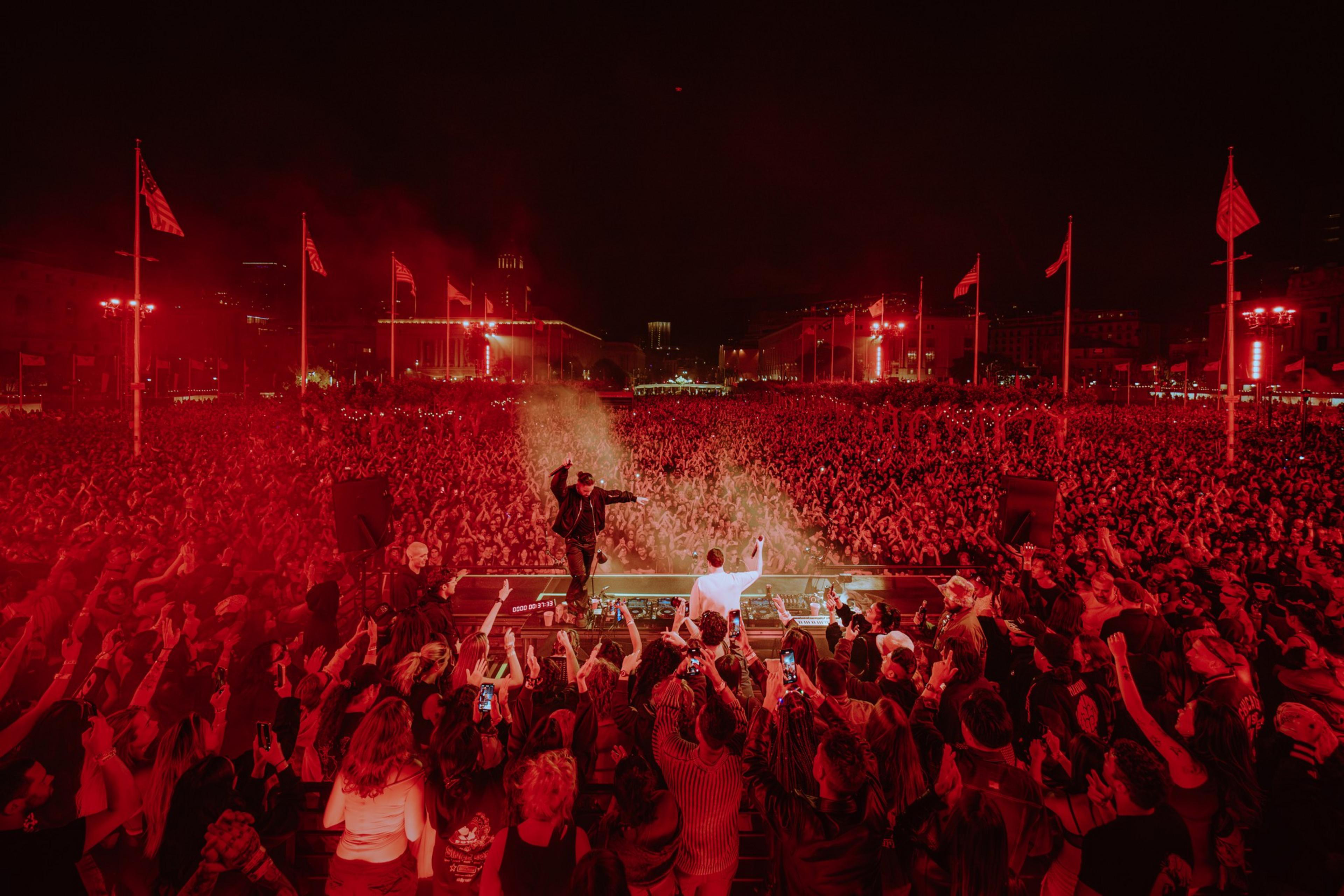 A musician performs on stage before a massive, energetic crowd at night, illuminated by red lighting and surrounded by waving flags.