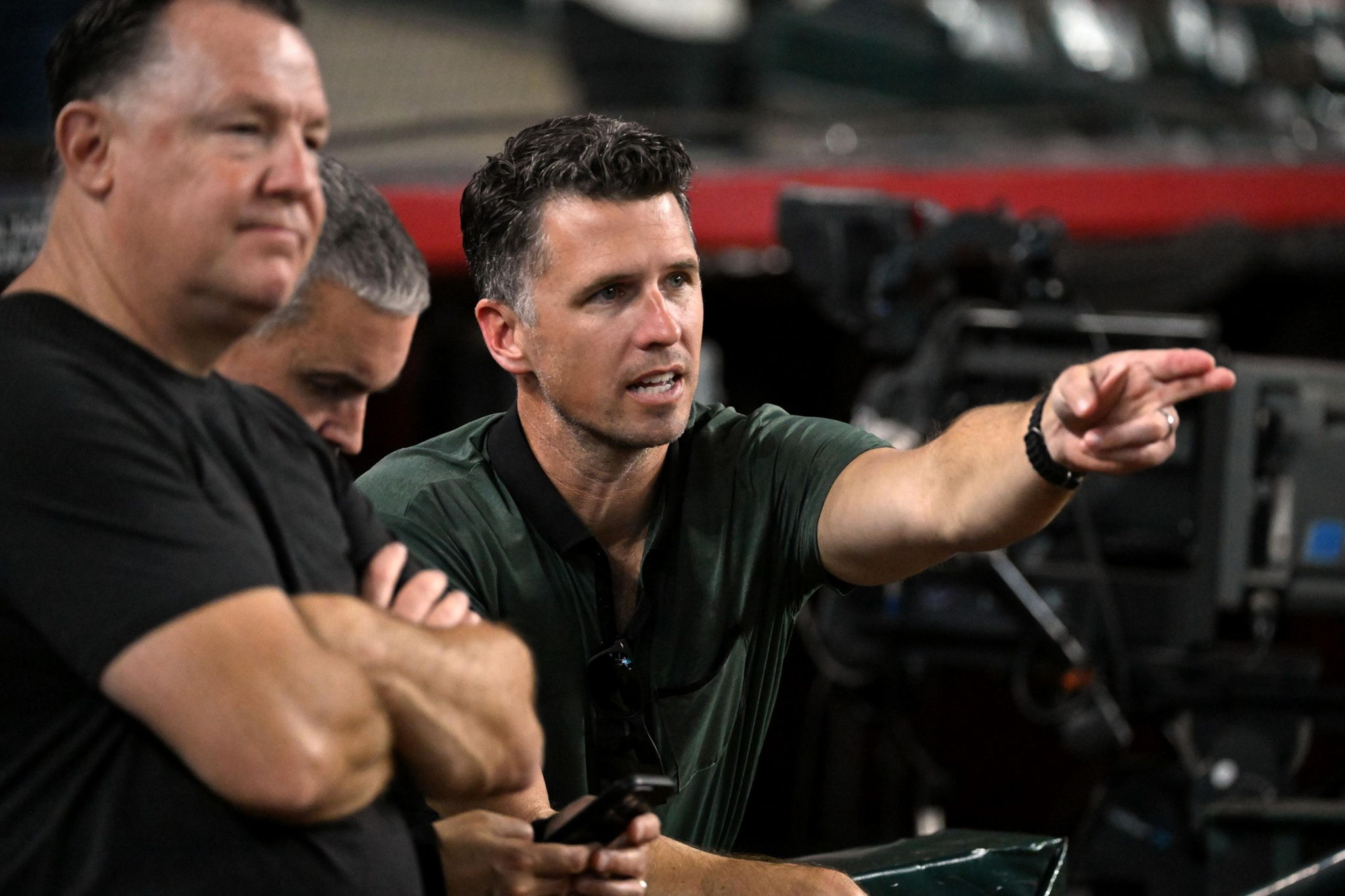 A man in a dark green shirt points intently while two men beside him sit with folded arms and looking away.