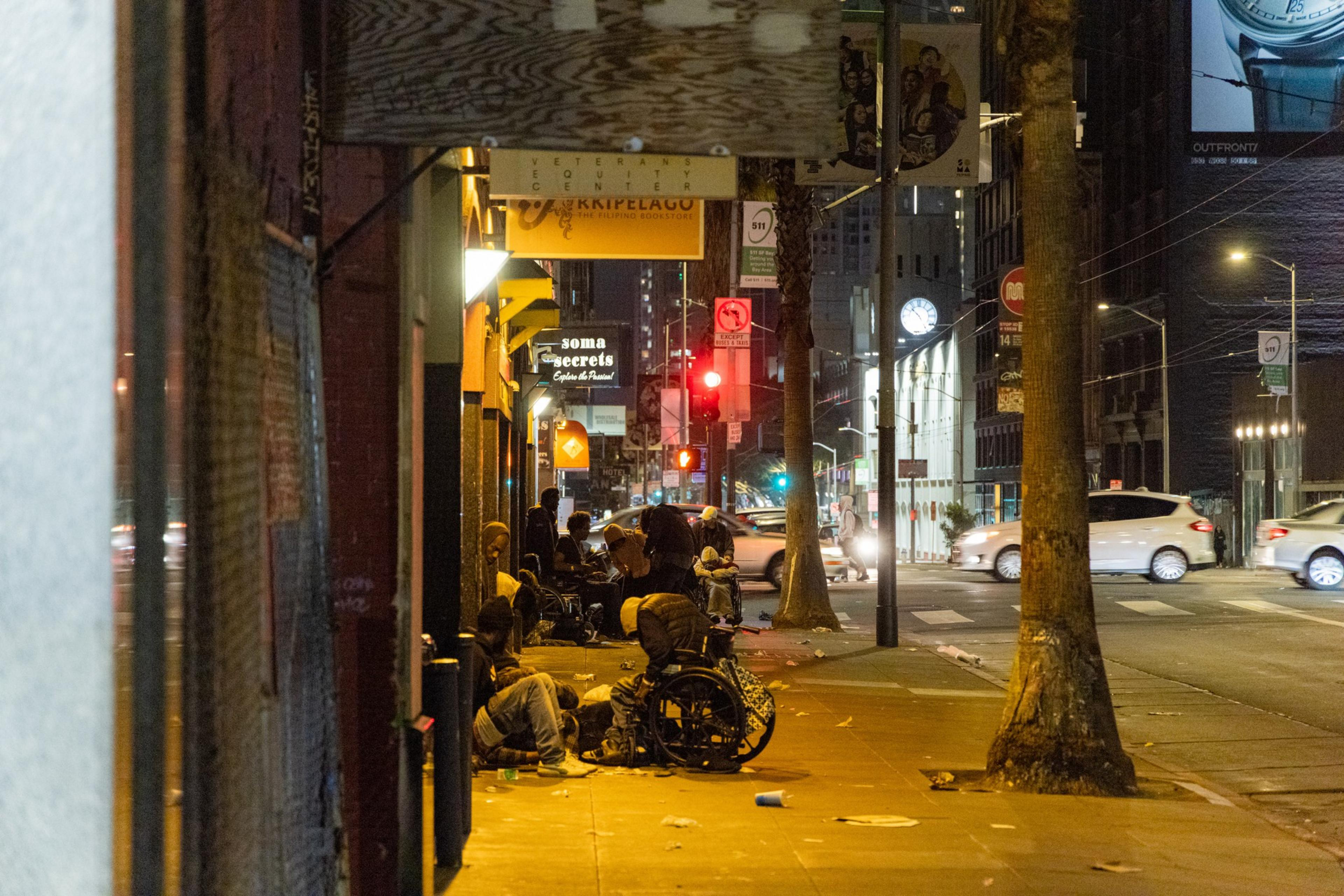At night on a city sidewalk, several people with wheelchairs congregate under streetlights, near storefronts. Cars and buildings with illuminated signs are in the background.